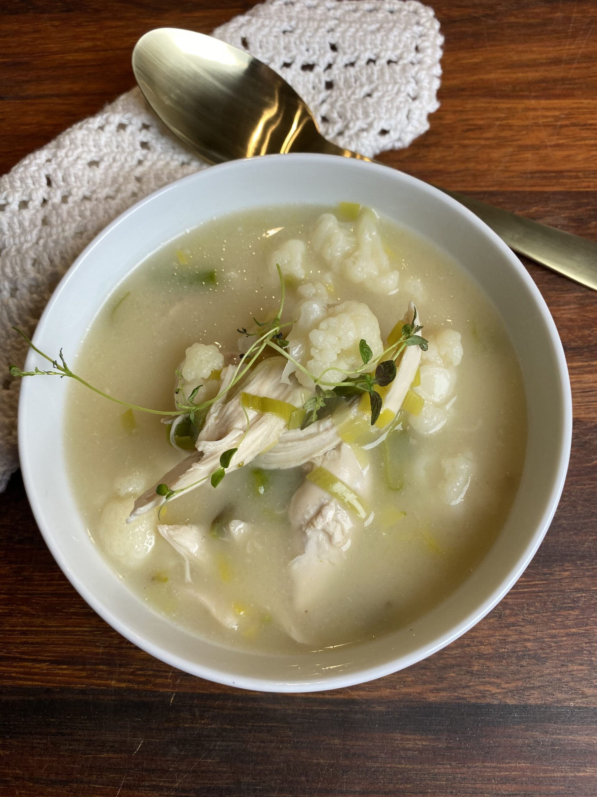 Kyllingesuppe med porrer, grønne asparges og blomkål i en hvid skål med en hæklet serviet ved siden af og en guld ske