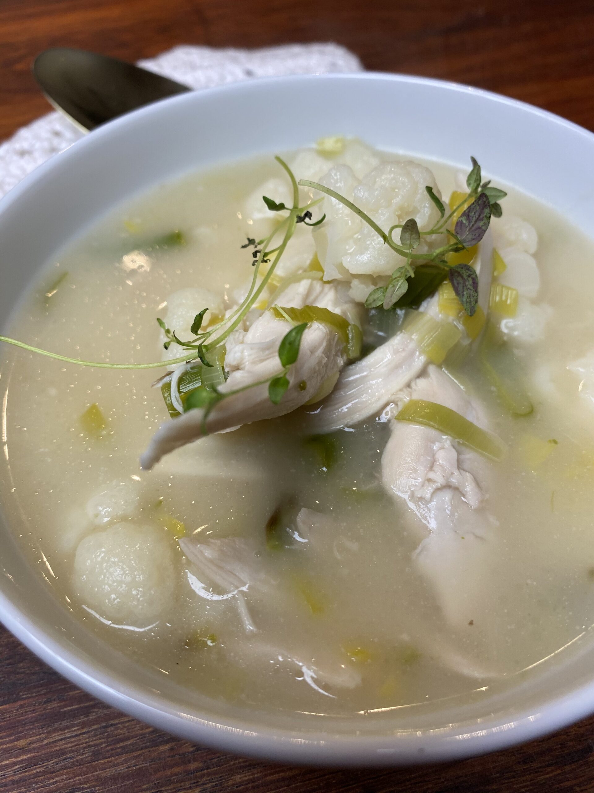 Kyllingesuppe med porrer, grønne asparges og blomkål i en hvid skål med en hæklet serviet og en guld ske ved siden af. Set tæt på.