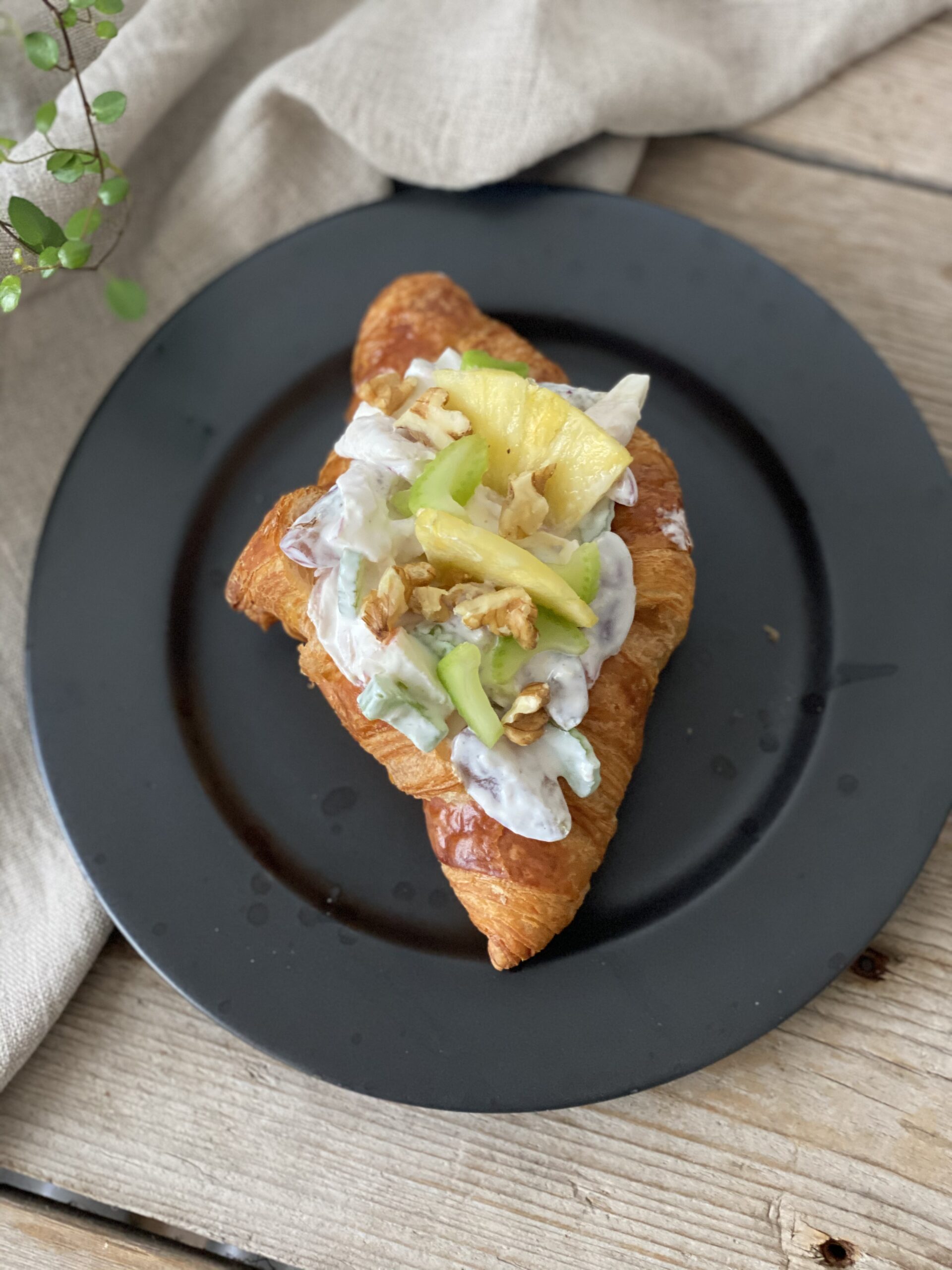 En croissant med kyllingesalat på en sort tallerken. Der er en hørserviet i baggrunden, det står på et bord lavet af træ og med en lille plante i det ene hjørne. Set oppefra.