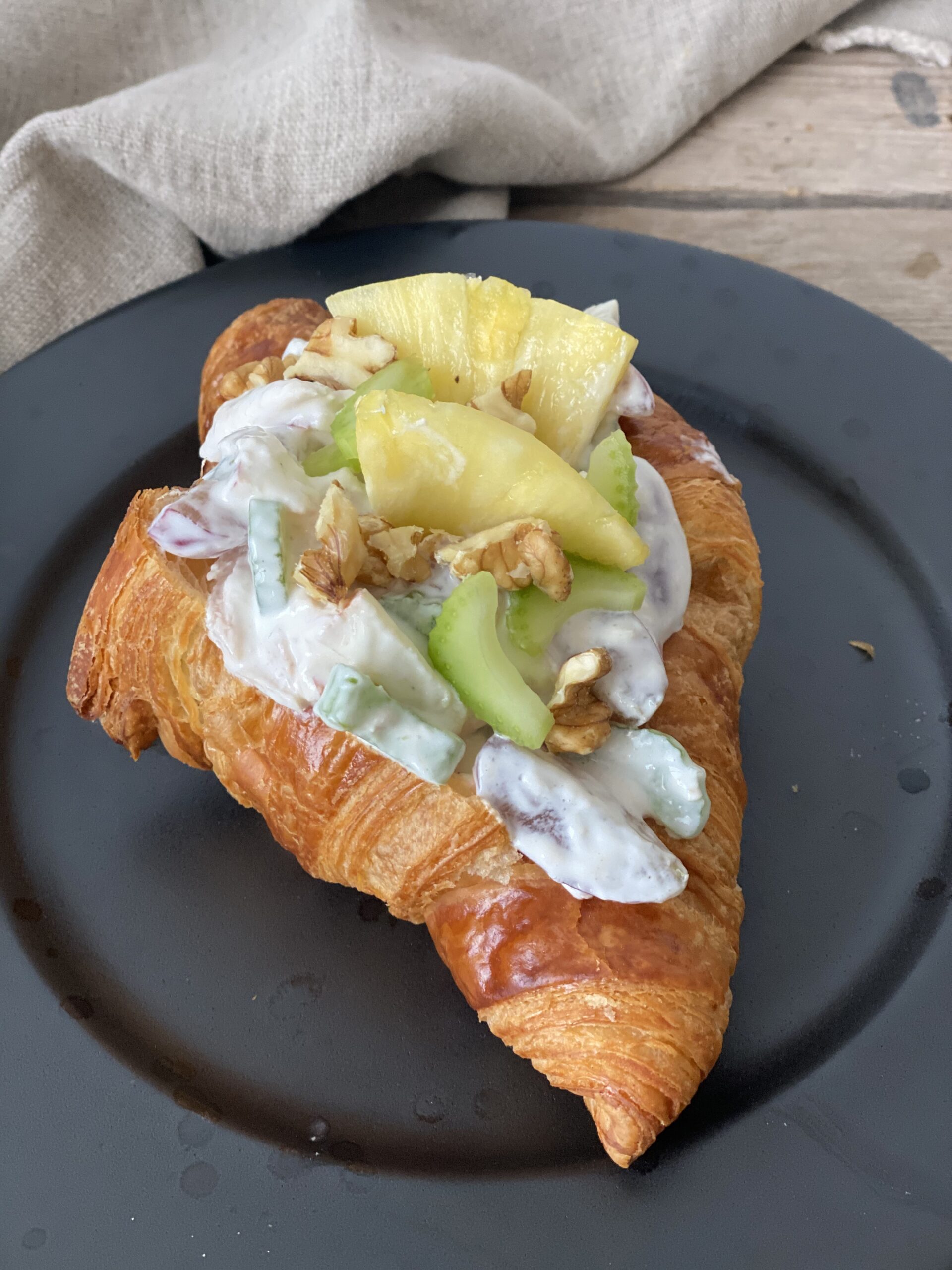 En croissant med kyllingesalat på en sort tallerken. Der er en hørserviet i baggrunden, det står på et bord lavet af træ og med en lille plante i det ene hjørne. Croissanten er set tæt på.