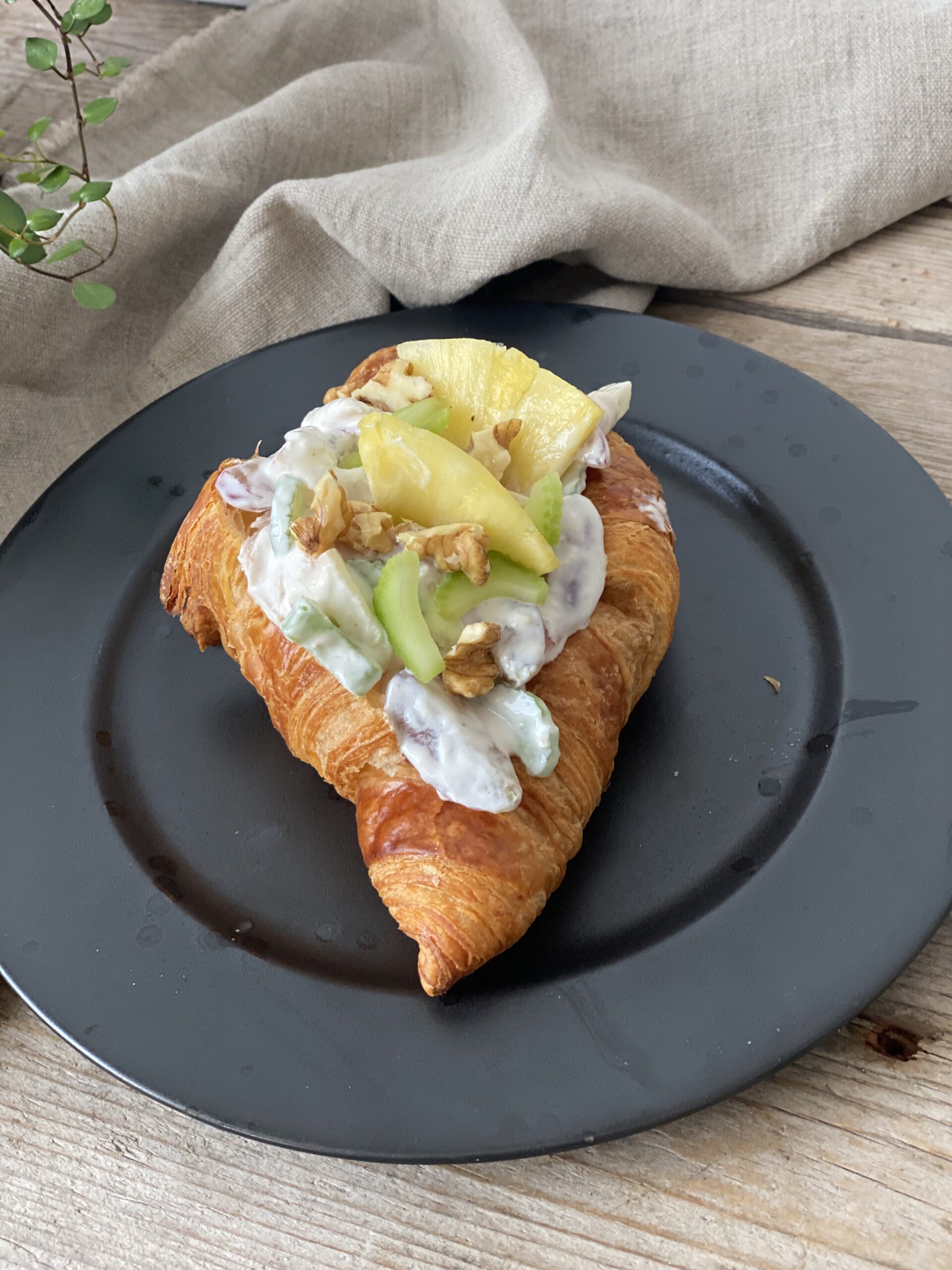 En croissant med kyllingesalat på en sort tallerken. Der er en hørserviet i baggrunden, det står på et bord lavet af træ og med en lille plante i det ene hjørne