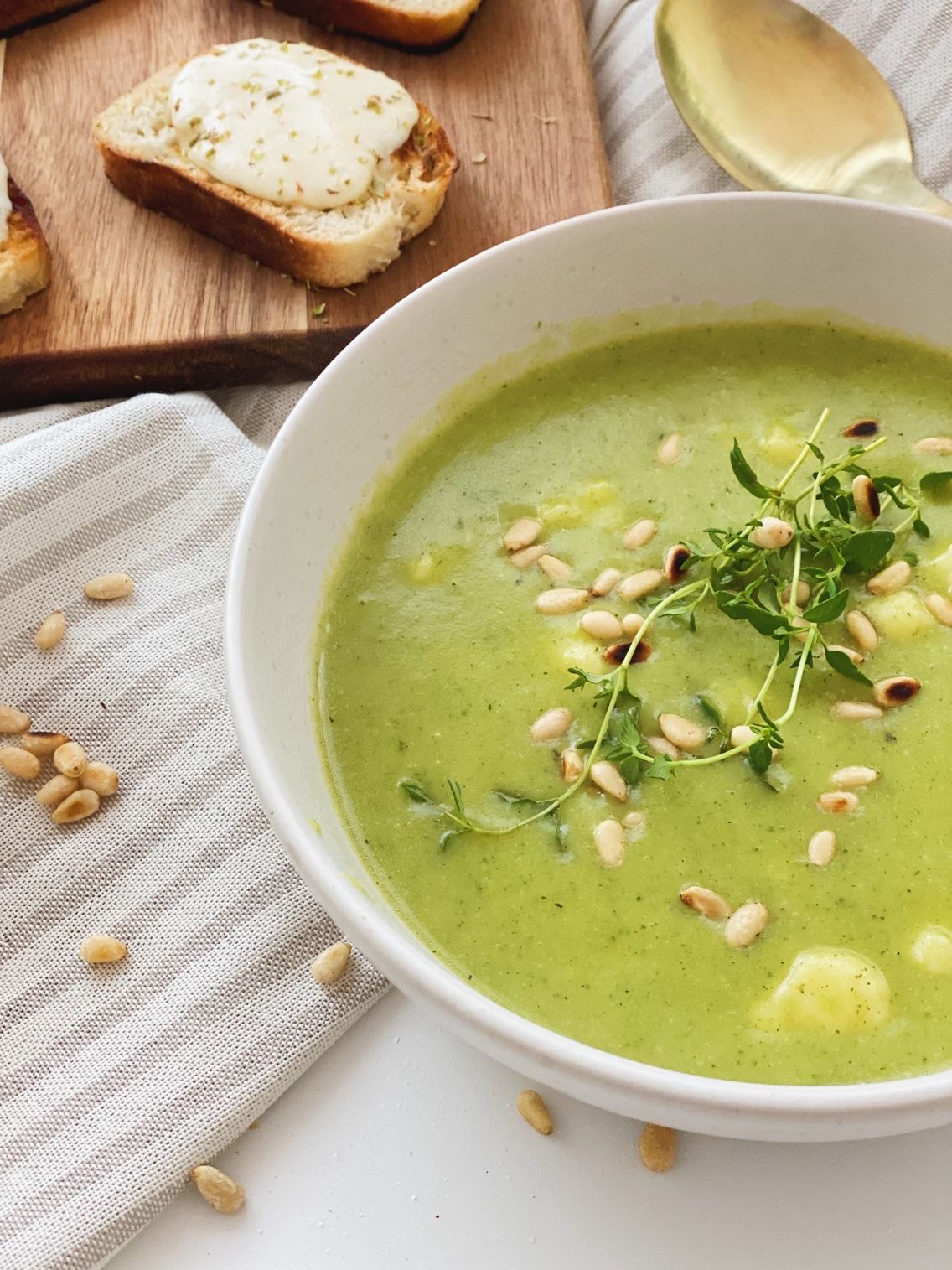 Et close up billede af broccolisuppe i en skål pyntet med ristede pinjekerner. Der ligger pinjekerner rundt på bordet, en ske ved siden af og ostebrød på et spækbræt ved siden af.
