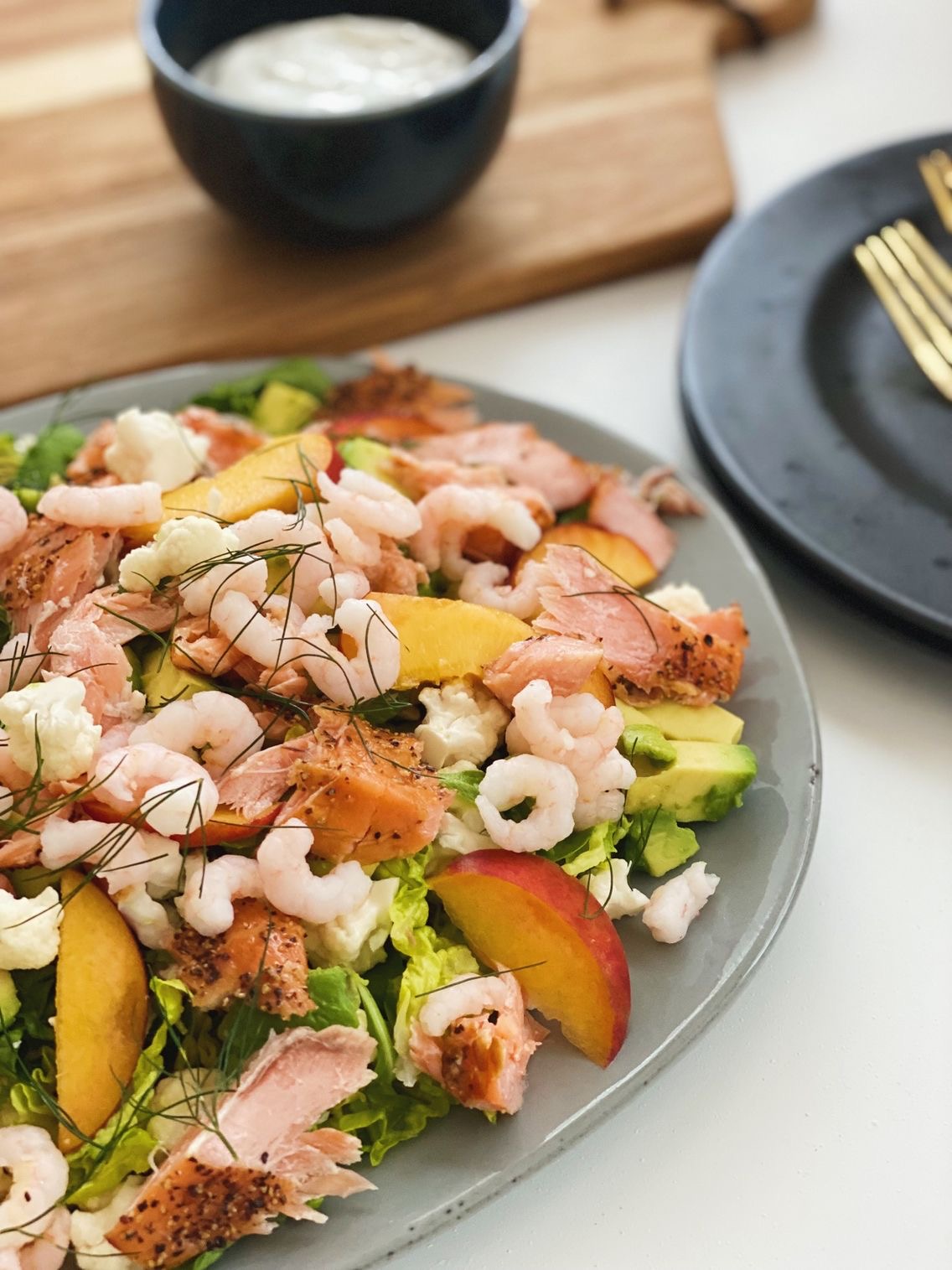 Måltidssalat med laks, rejer og ferskner på et gråt fad set tæt på med en skål med cremefraichedressing i baggrunden som er i en sort skål. Der står til højre tallerkner som der ligger bestik på