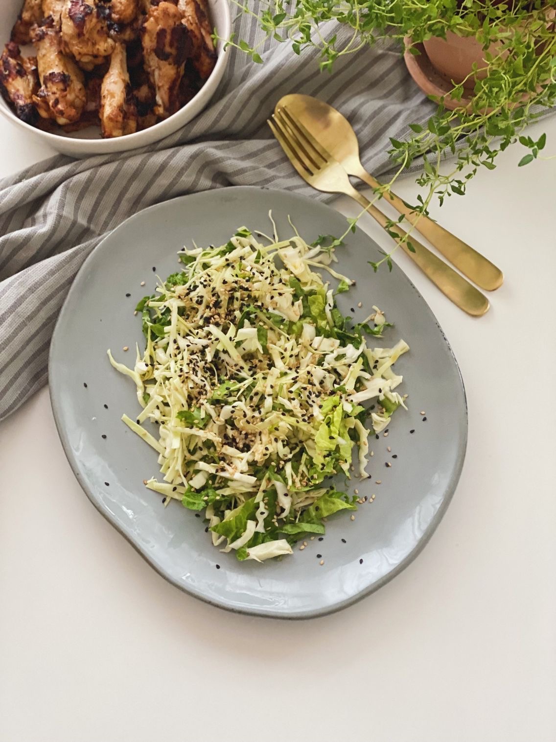 Spidskålsalat med ingefærdressing på et gråt fad set oppefra med kyllingevinger i baggrunden og frisk timian i en lerpotte