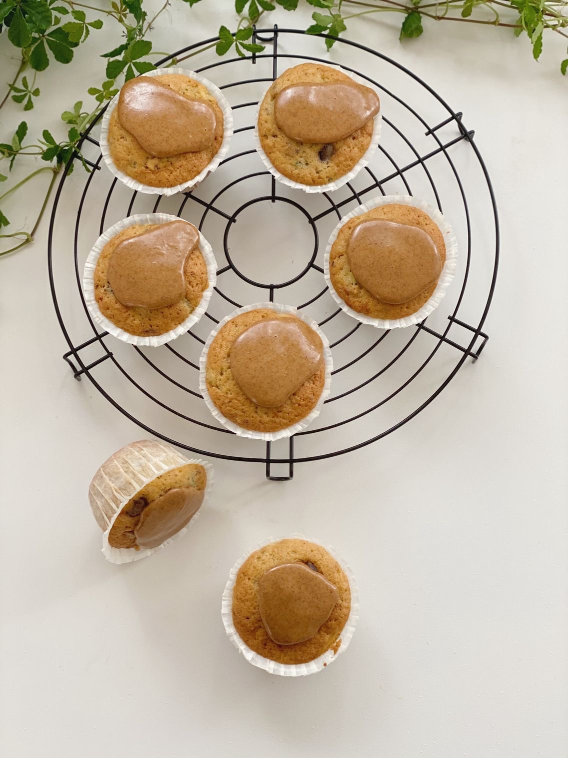 Squashmuffins på en rund bagerist med 5 muffins på med kanelglasur set oppefra. Ved siden af risten ligger der en muffins på siden og en står oprejst. I baggrunden kan man se nogle grønne blade fra en plante.