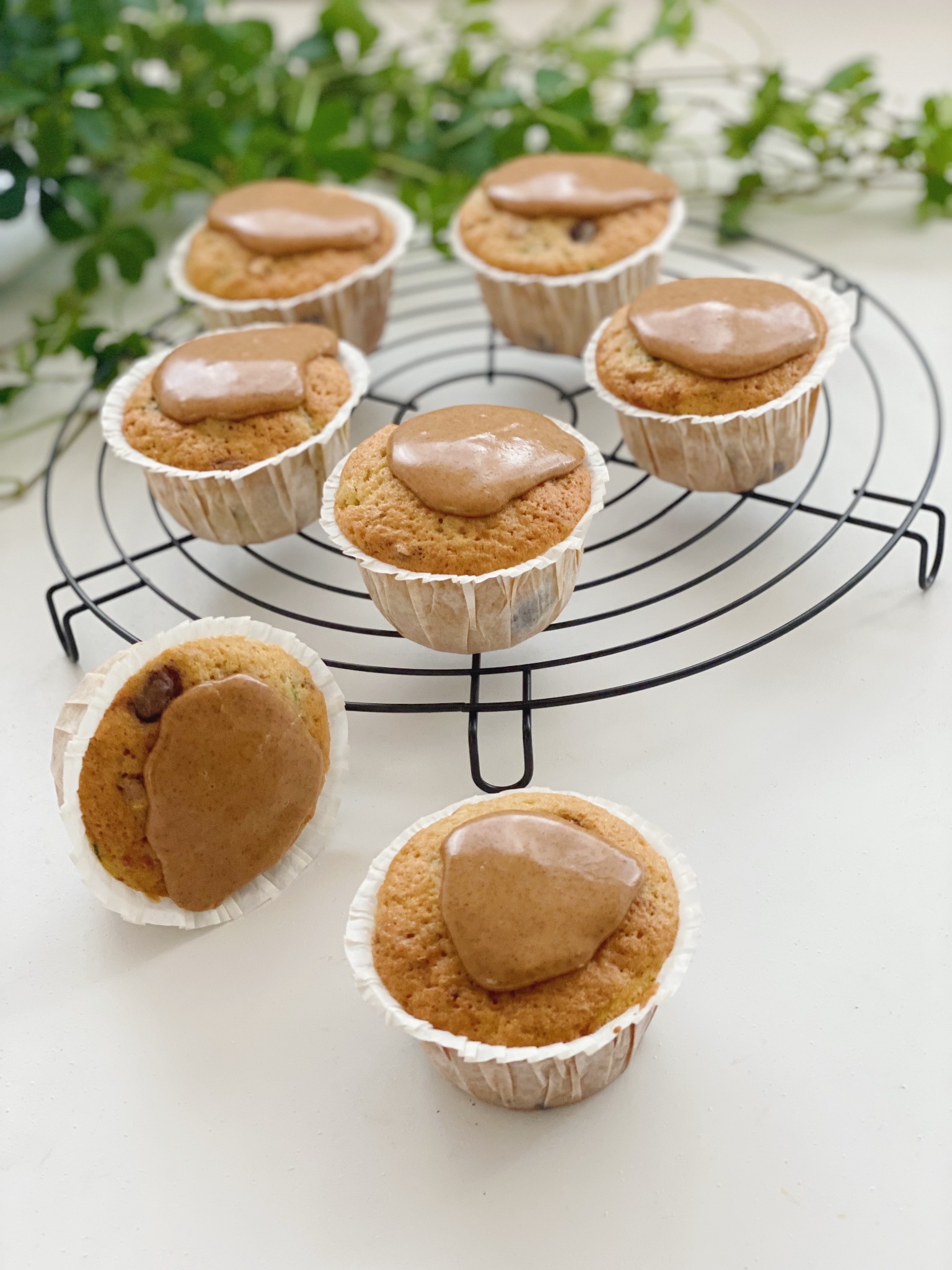 Samme billede som det forige med 5 squashmuffins på en rund bagerist med kanelglasur. Ved siden af risten ligger der en muffins på siden og en står oprejst. I baggrunden kan man se nogle grønne blade fra en plante. Det hele er ses mere direkte ind på billedet og tættere på.