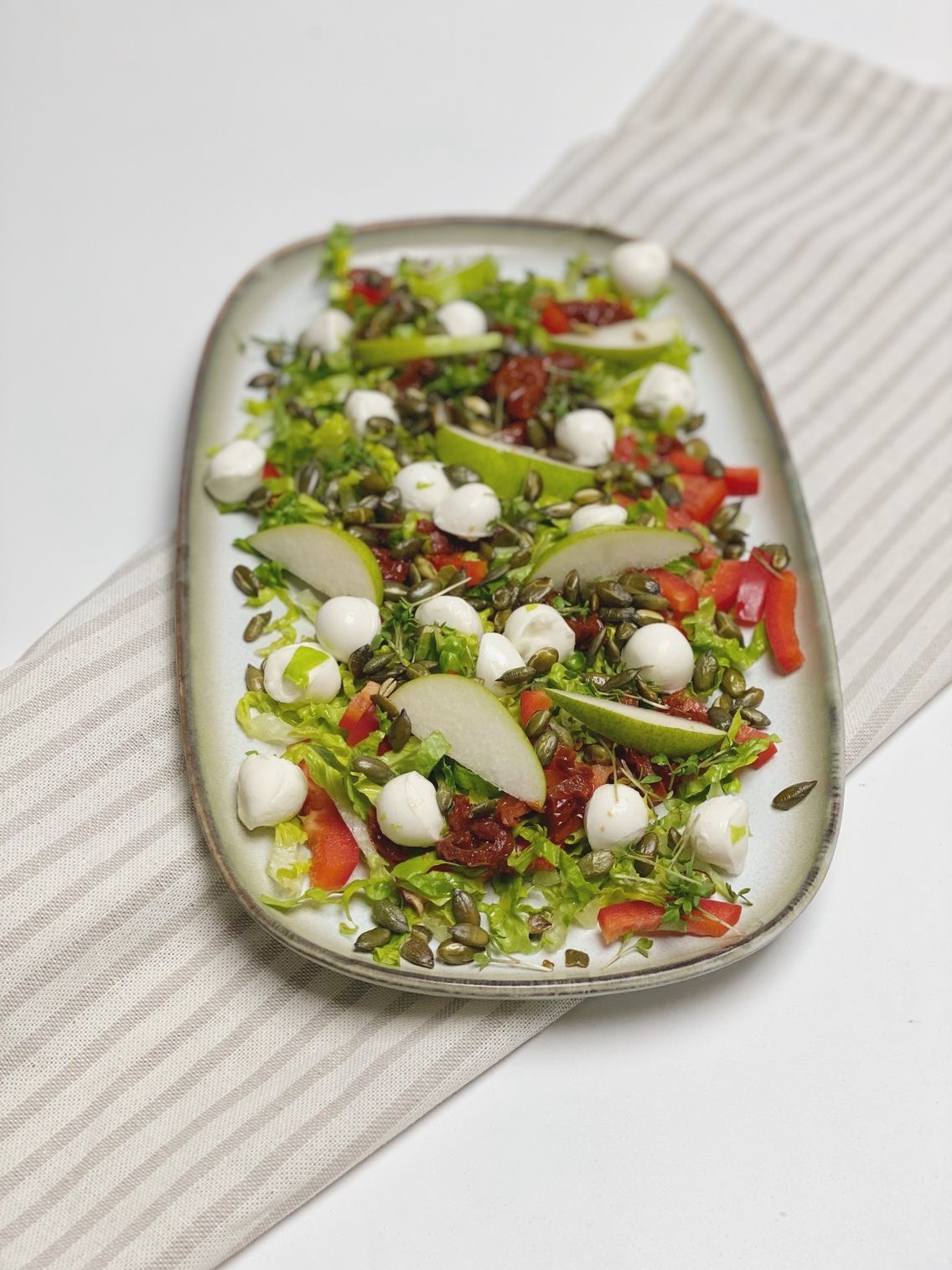 Salat med pære og mozzarella som er anrettet på et aflangt fad som er placeret oven på en stribet serviet. Set skråt ind oppefra. Man kan se hele fadet.