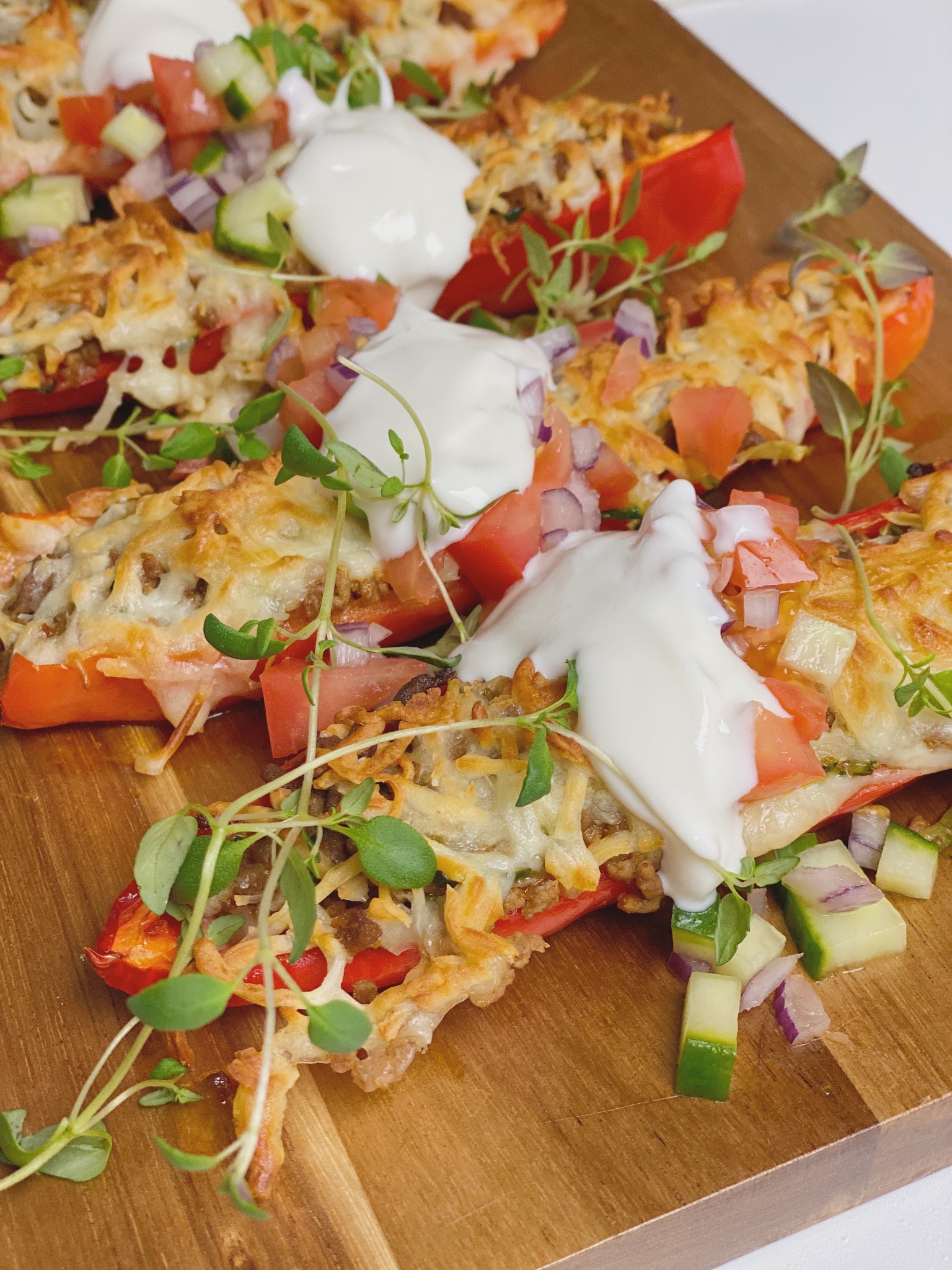 Fyldte snackpeber med tomatsalat og en klat cremefraiche på. Man kan se fire halve snackpebre med fyld på et skærebræt af træ. Billedet er et close up billede, hvor du ser dem meget tæt på.