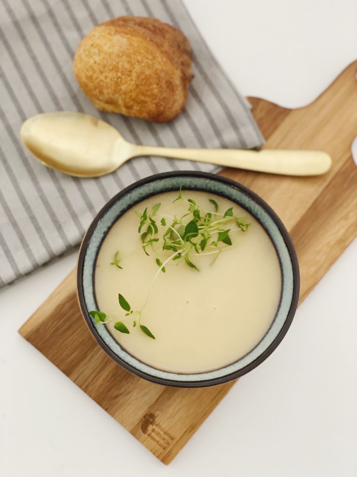 Kartoffel selleri suppe set oppe fra. Suppen er i en blå skål og der er en kvist timian oven på suppen. Suppen står på et lille træskærebræt og der ligger en guldske på en stribet serviet og et kuvertbrød