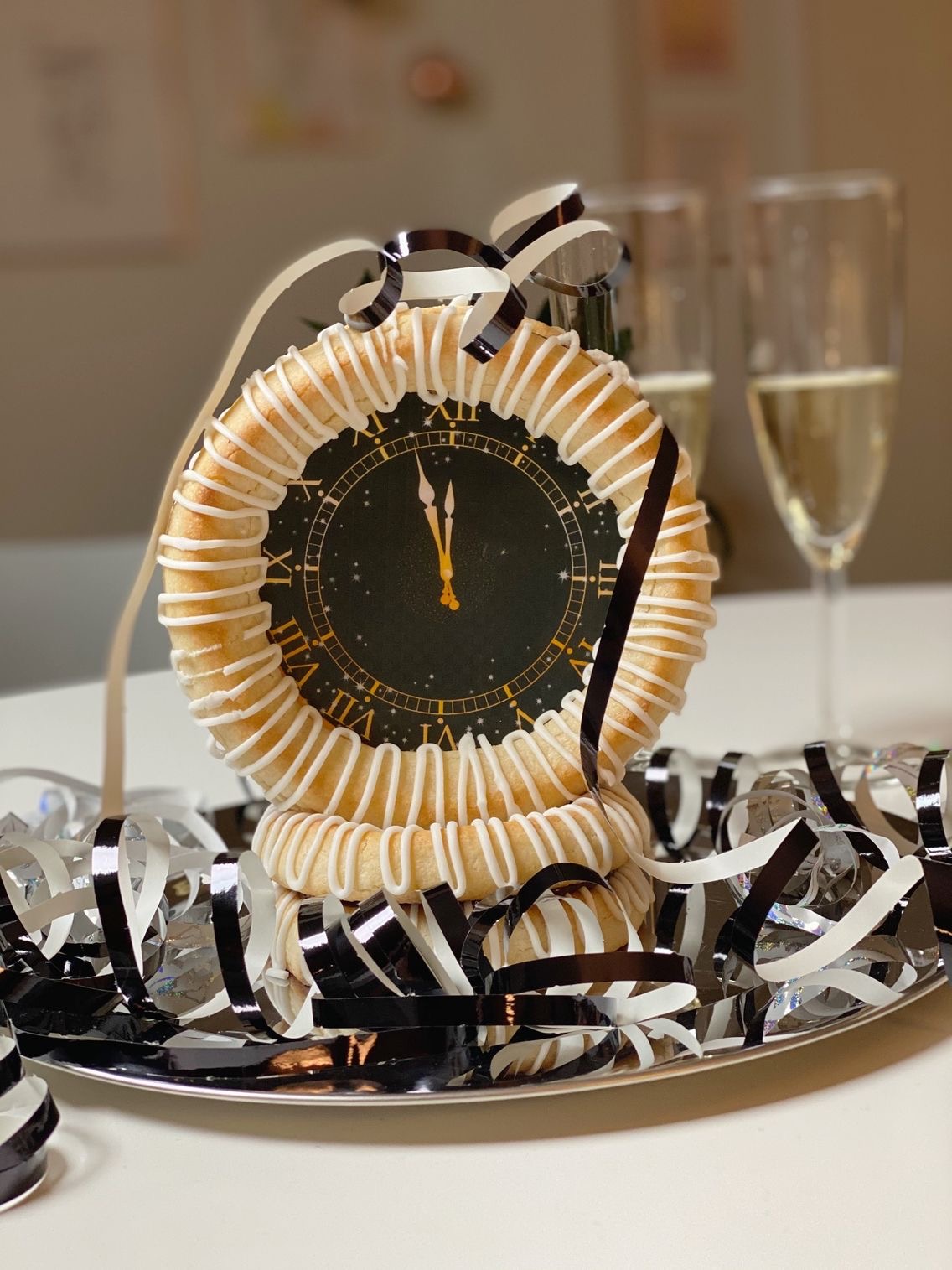 Nytårsur af kransekage med som står på et sølvfad omgivet af sorte serpentiner. Der står to glas champagne bagved