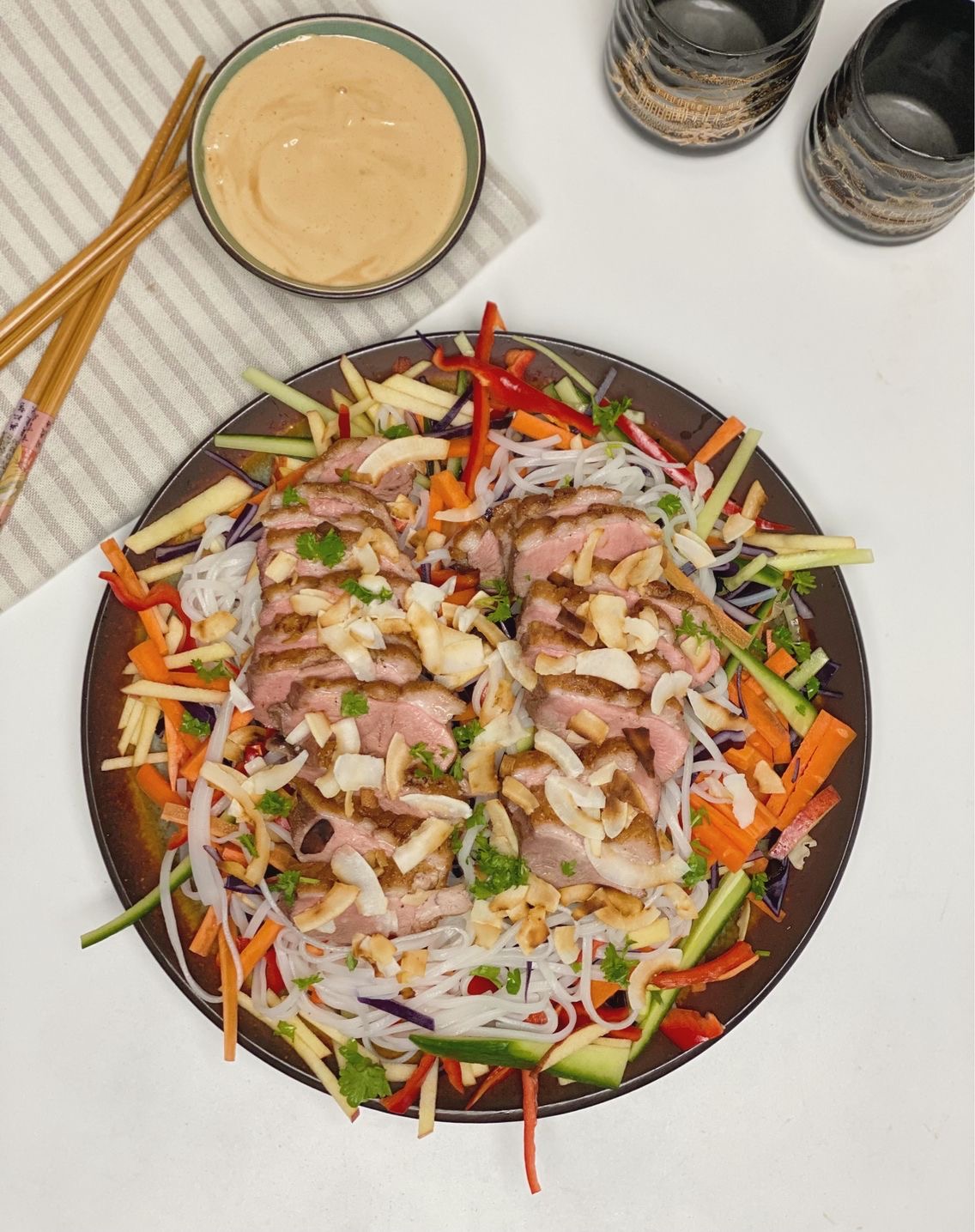 Asiatisk inspireret måltidssalat med andebryst set oppefra med en stribet serviet ved siden af, hvor der ligger to sæt spisepinde og en skål med peanutbutter dip. Til den anden side står der to kinesiske kopper som er sorte med guld skrift på.