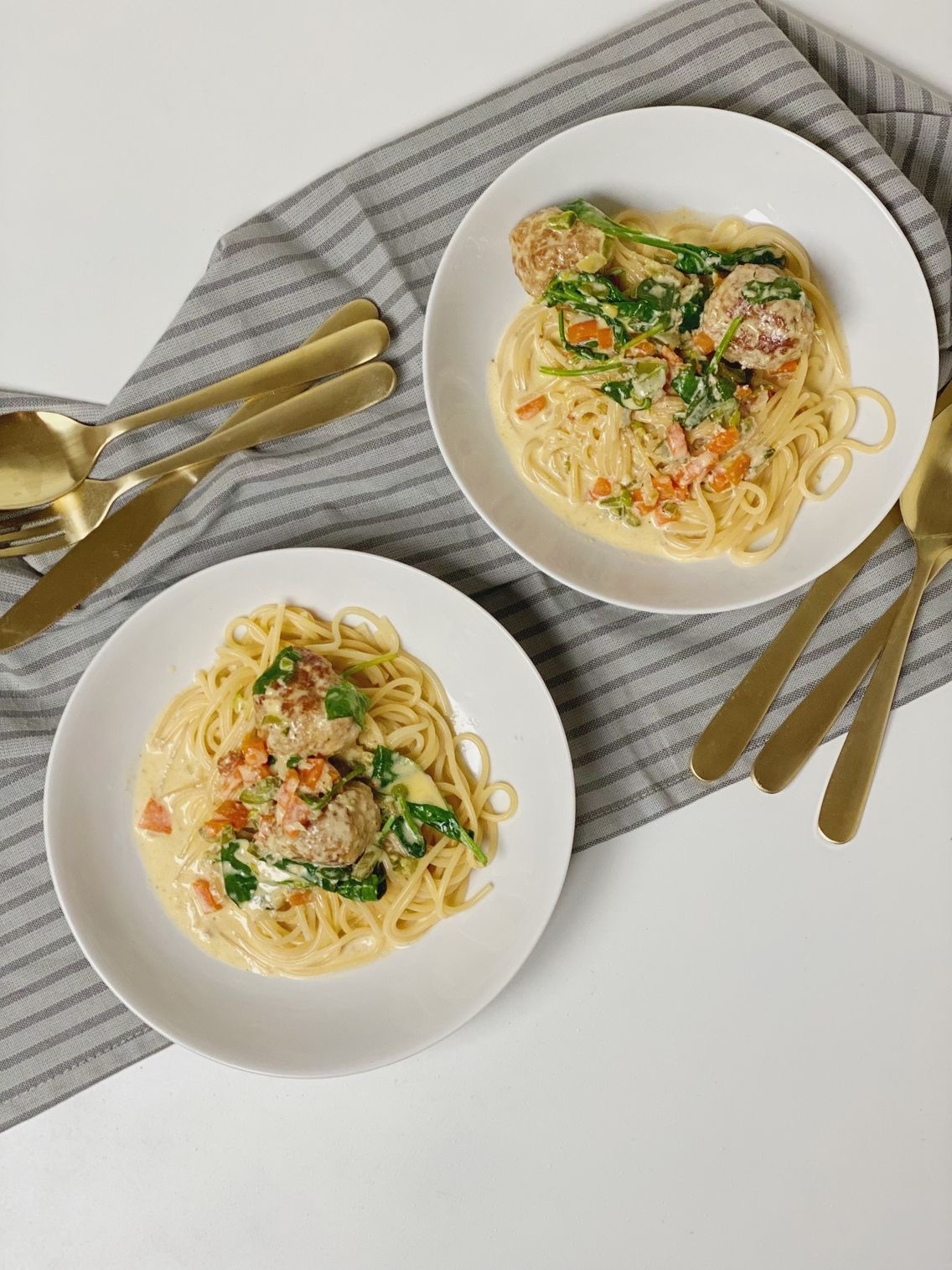 Spicy kødboller anrettet i to hvide dybe tallerkner med spaghetti og to kødboller i hver og flødesovsen hældt ud over.
Billedet er taget oppe fra så man ser begge tallekener i fuld størrelse og med bestik ved siden af. Det hele står oven på en stribet serviet i grå nuancer