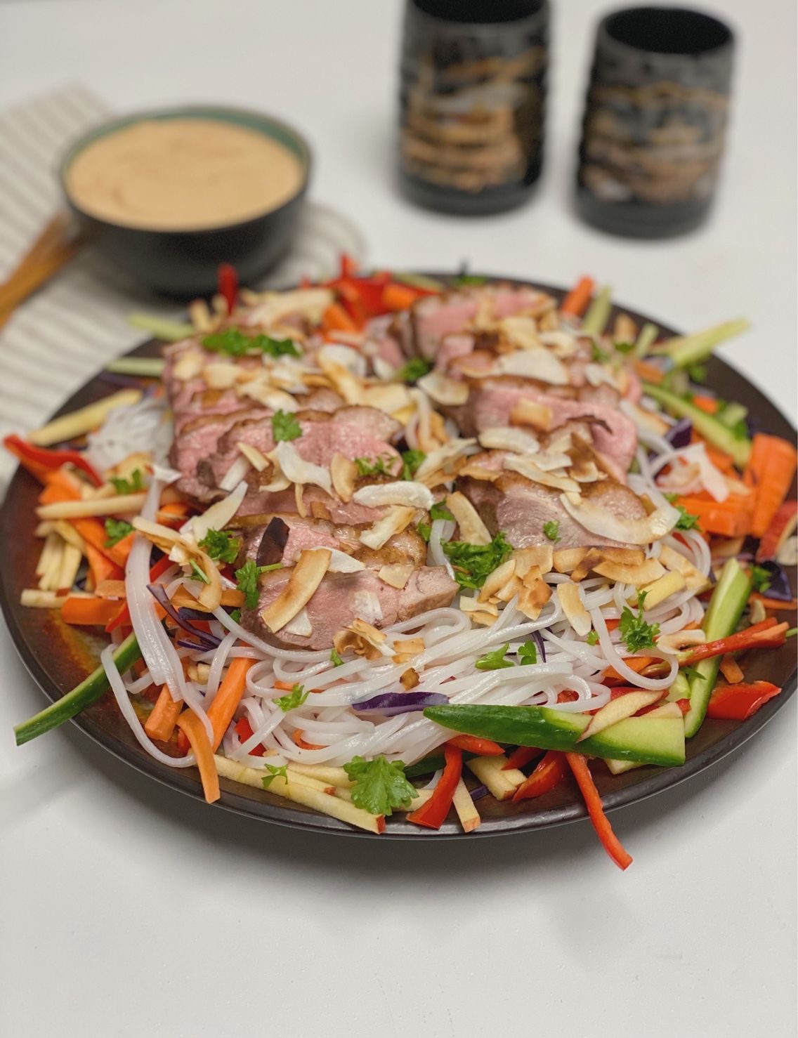 Asiatisk inspireret måltidssalat med andebryst set vandret ind med en stribet serviet ved siden af, hvor der ligger to sæt spisepinde og en skål med peanutbutter dip. Til den anden side står der to kinesiske kopper som er sorte med guld skrift på
