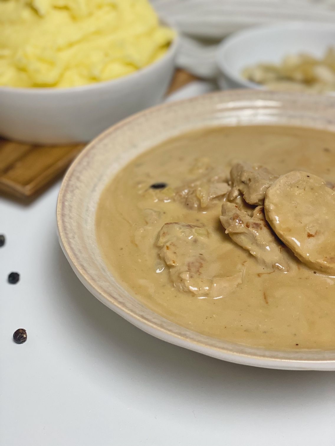 Svinemørbrad med enebær i ginsauce set tæt på, hvor du kun ser halvdelen af skålen. I baggrunden kan du se en skål med kartoffelmos og nogle syltede asier. På bordet ligger der nogle tørrede enebær