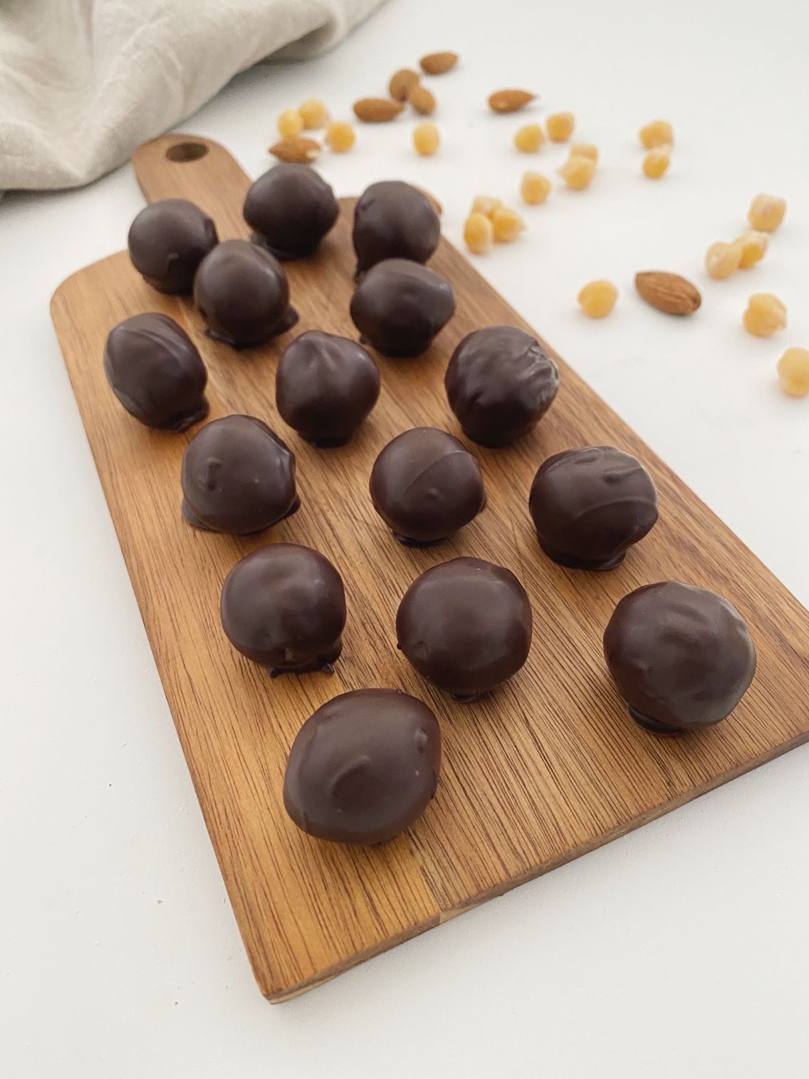 Marcipankugler overtrukne med chokolade, der ligger på et træ skærebræt. Man kan i baggrunden se nogle kikærter og mandler