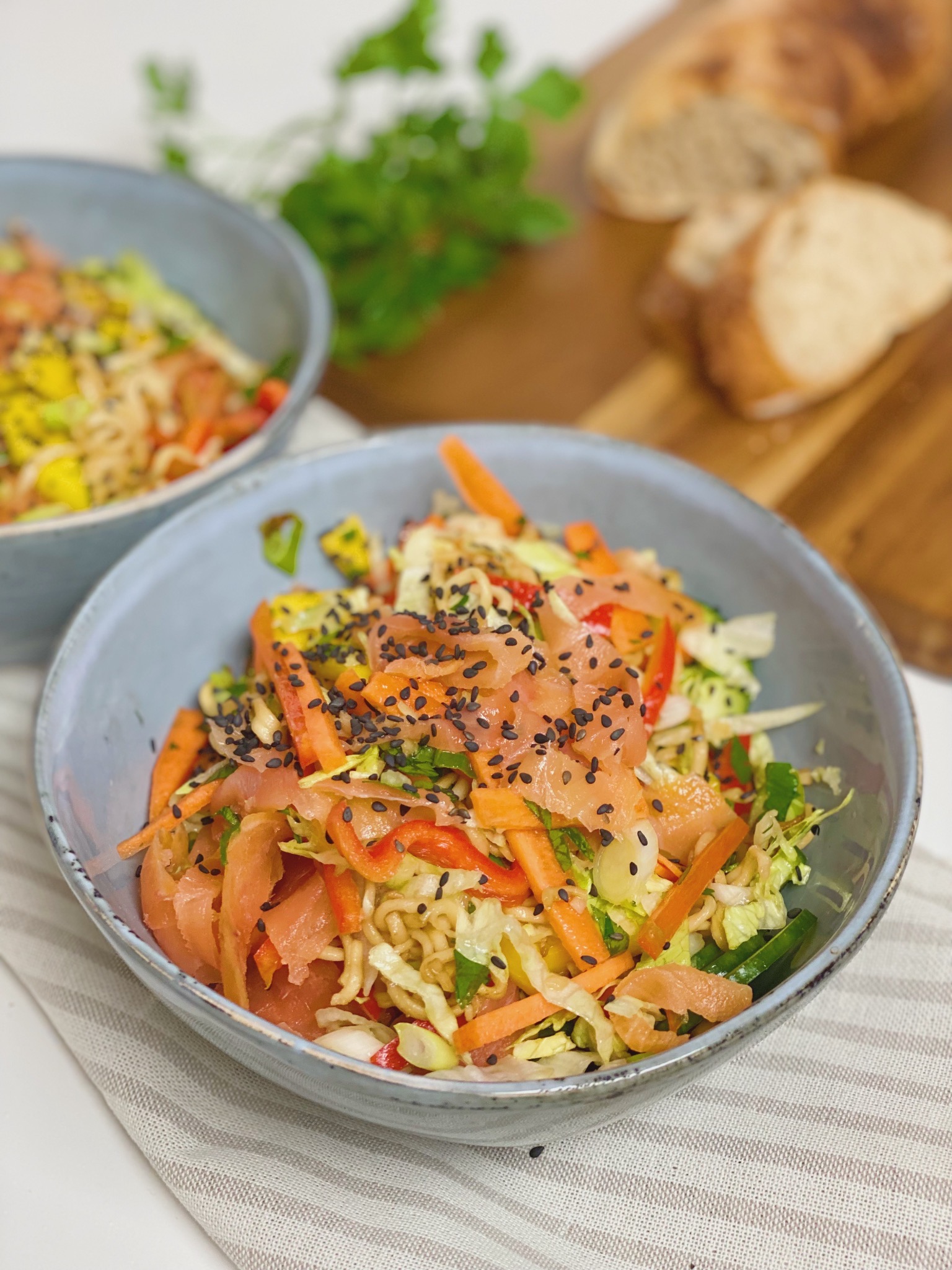 to skåle med salatbowl med nudler og koldrøget laks, hvor der er en spækbræt i baggrunden med hjemmebagt flutes på. Den ene skål står tæt på og den anden i baggrunden
