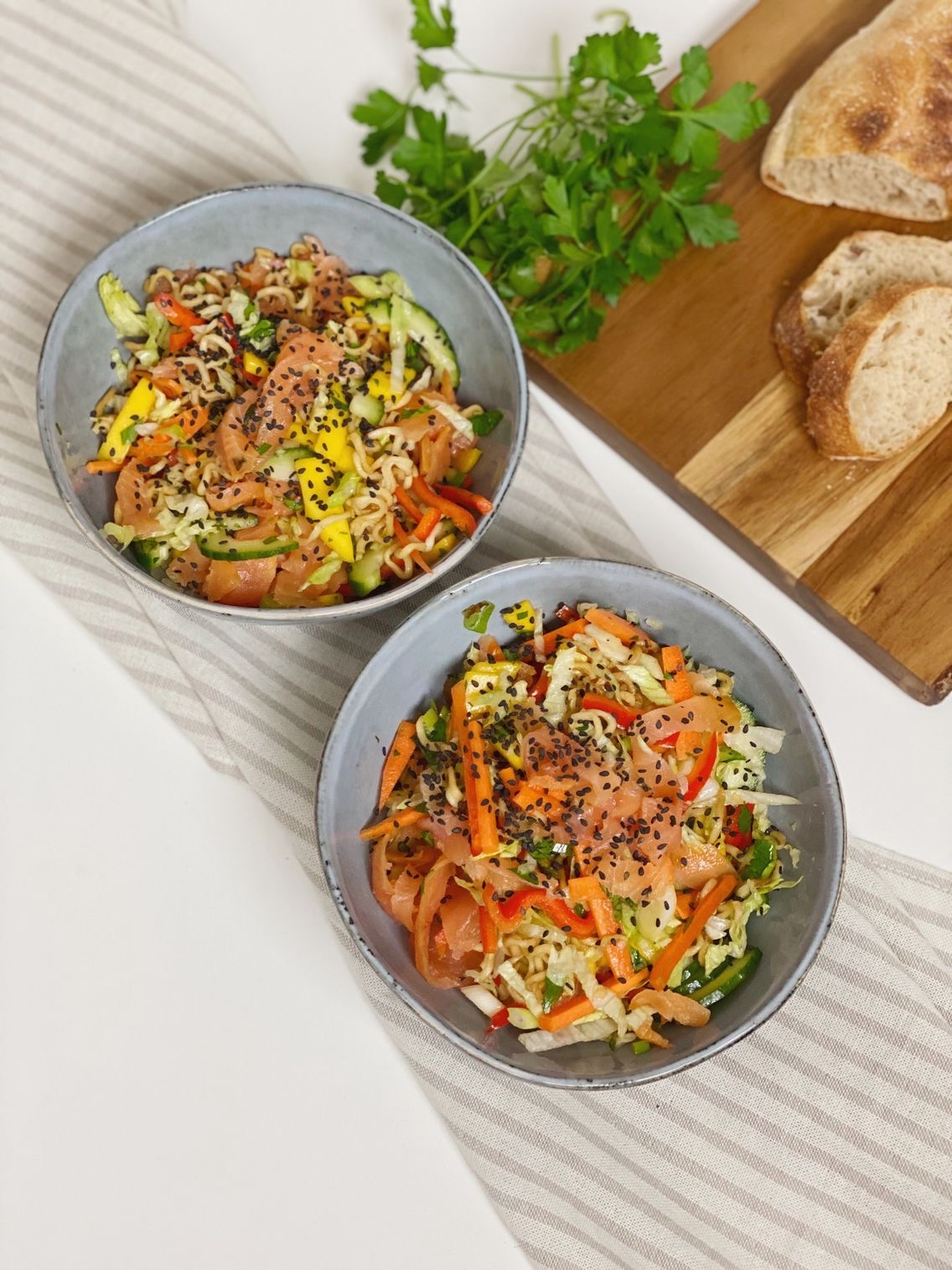 to skåle med salatbowl med nudler og koldrøget laks, hvor der er en spækbræt i baggrunden med hjemmebagt flutes på. Det hele er set oppefra