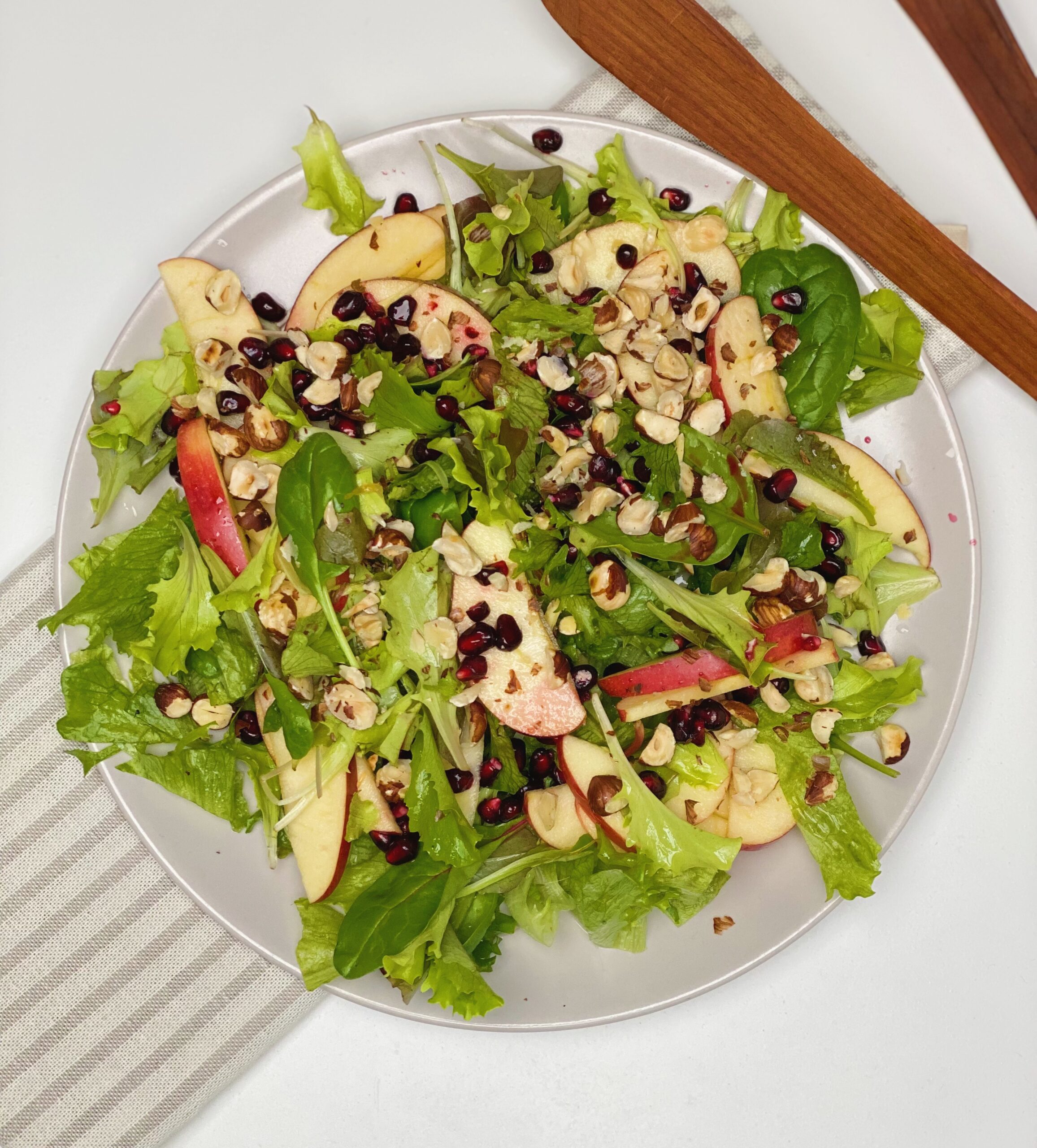 Salat med appelsinvinaigrette set oppefra hvor man kan se hele tallerkenen. I baggrunden ligger der en salattang og tallerkenen står oven på en stribet serviet.