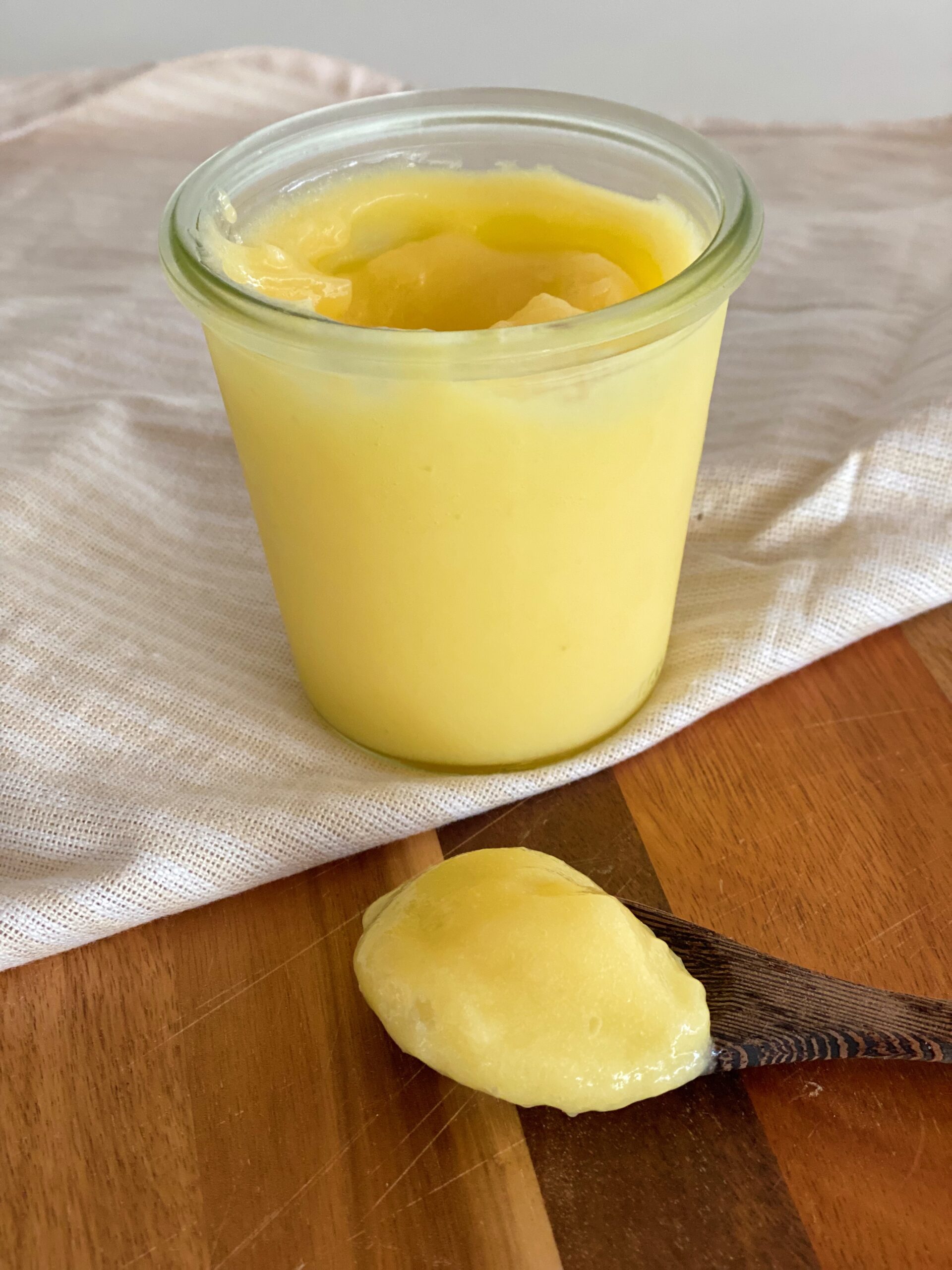 Lemoncurd i en glasskål som står på en stofserviet ovenpå et træ skærebræt- FOran ligger der en træske med lemoncurd på.