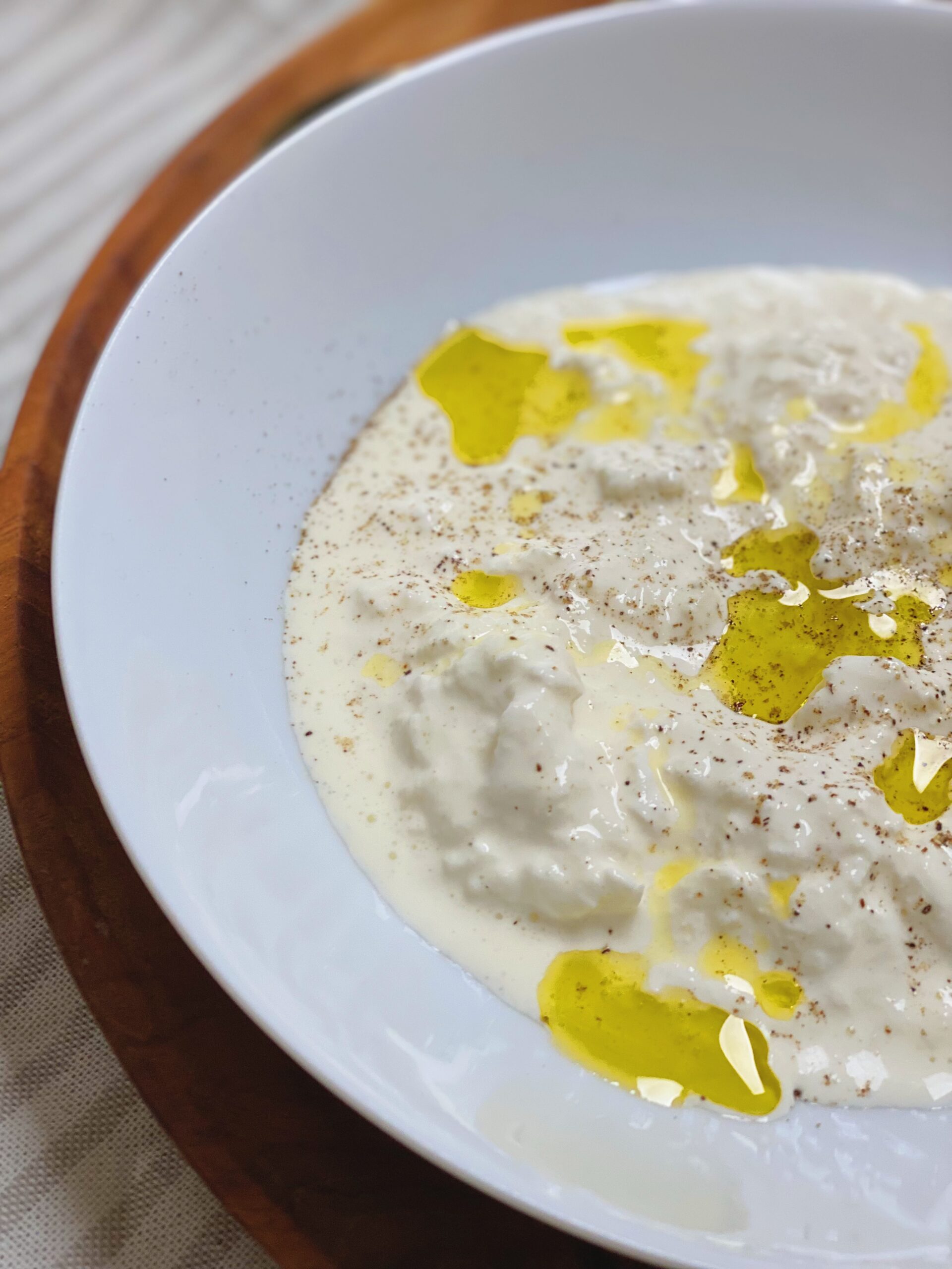Stracciatella i en hvid dyb tallerken med olivenolie på og friskkværnet peber. Tallerkenen står på et træfad. Du ser kun halvdelen af tallerkenen