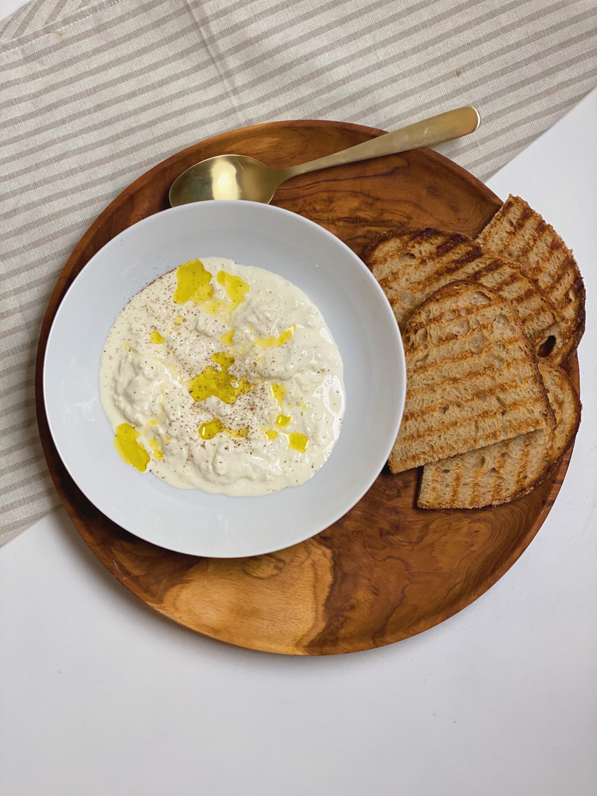 Stracciatella i en hvid dyb tallerken med olivenolie på og friskkværnet peber. Tallerkenen står på et træfad med 4 skiver ristet brød