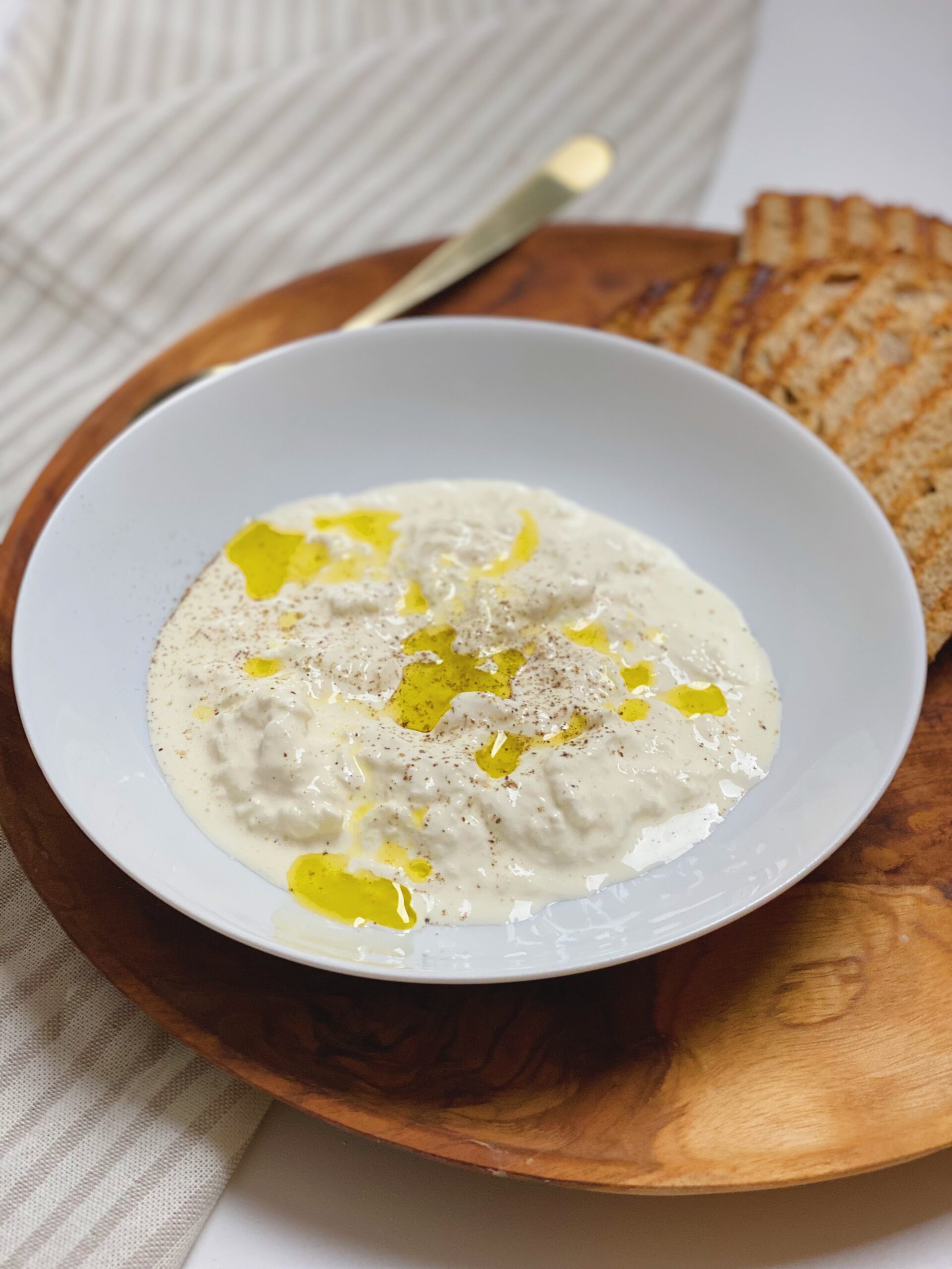 Stracciatella i en hvid dyb tallerken med olivenolie på og friskkværnet peber. Tallerkenen står på et træfad med 4 skiver ristet brød. Det hele er set tæt på