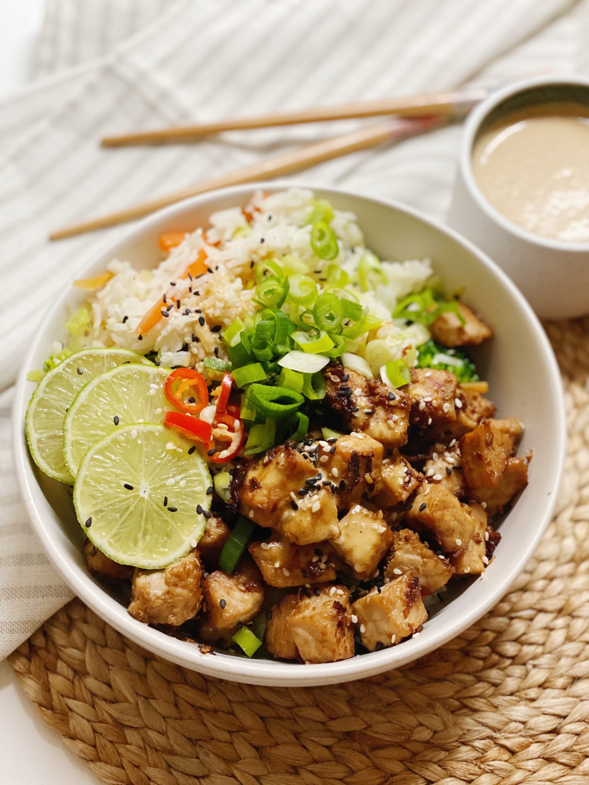 Asiatisk inspireret ret med marineret svinekød i en hvid skål set tæt på. der står en skål med peanutbutter sauce ved siden af. Der ligger en serviet og nogle spisepinde og skålene står oven på en siv dækkeserviet
