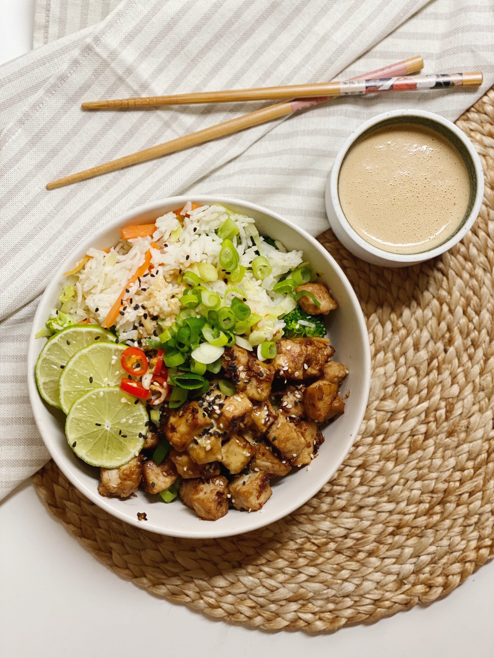 Asiatisk inspireret ret med marineret svinekød i en hvid skål set oppefra. der står en skål med peanutbutter sauce ved siden af. Der ligger en serviet og nogle spisepinde og skålene står oven på en siv dækkeserviet