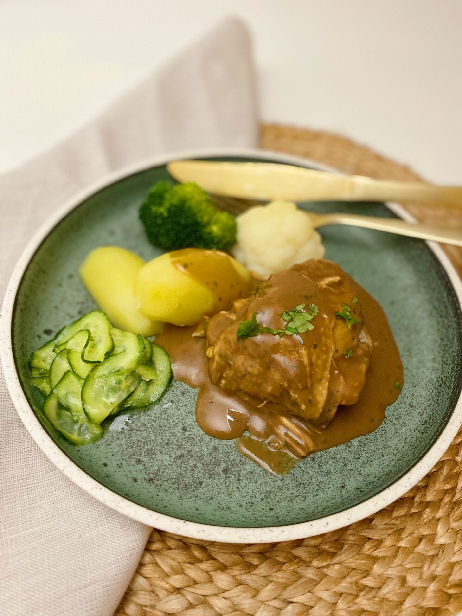 Den grydestegte kylling med sovs på serveret på en grøn tallerken. Ved siden af kyllingen ligger der kogte kartofler, blomkål og broccoli og agurkesalat.