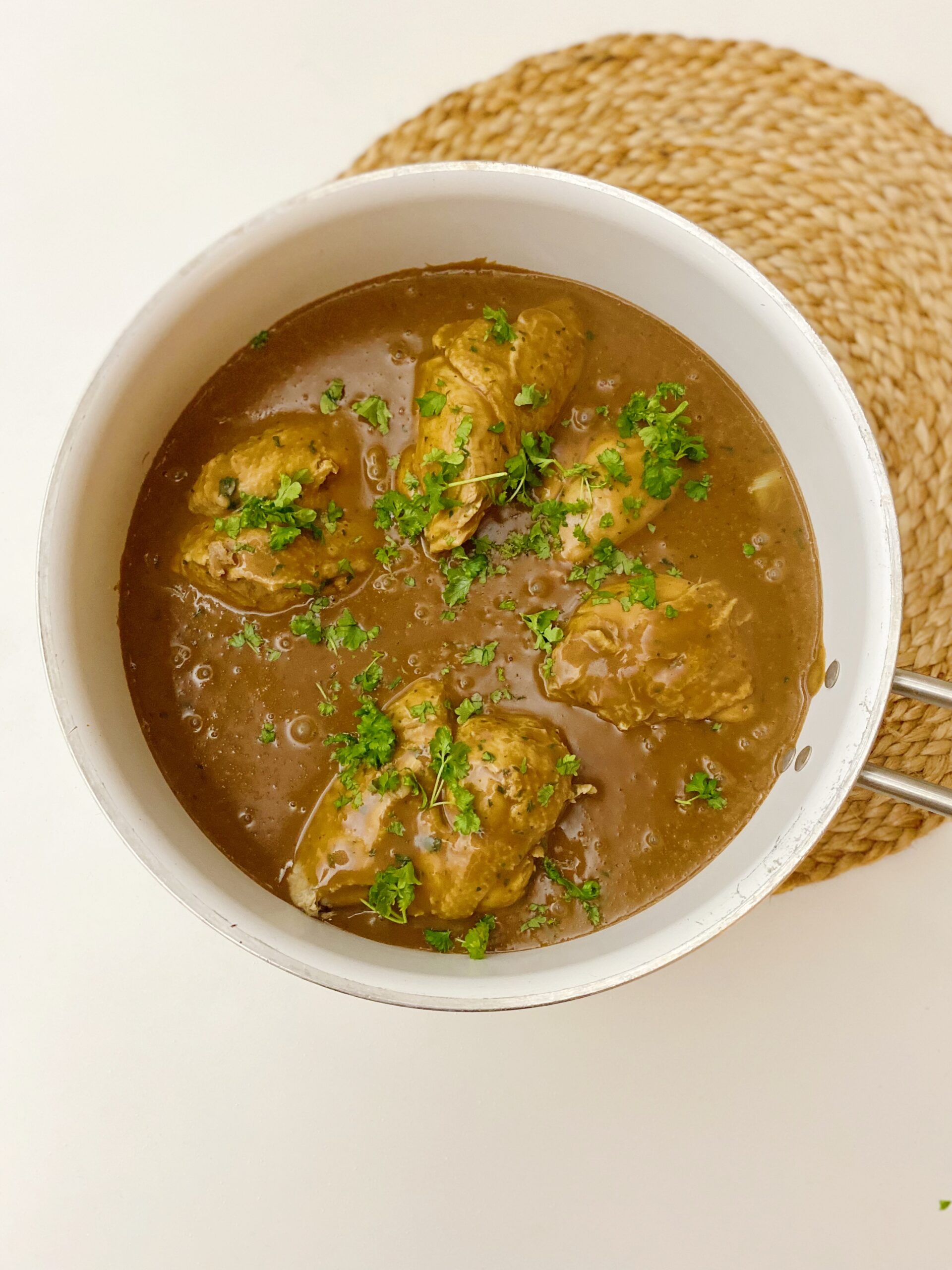 Grydestegt kylling i en dejlig lysebrun sauce pyntet med hakket persille. Det er i en hvid sautepande som står ovenpå en bambus dækkeserviet.