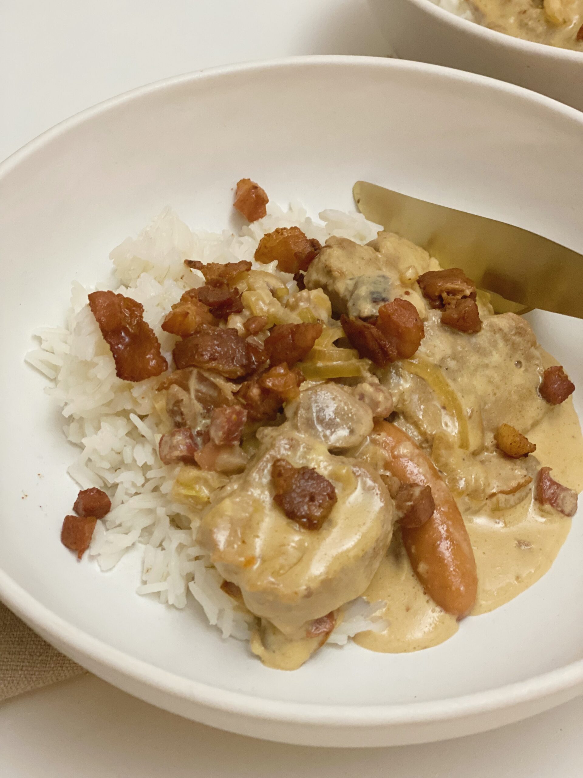 En portion mørbrad ala creme serveret i en dybe tallerkner med kogte ris til. Der er bestik af "guld" som ligger i tallerkenen. Set tæt på.