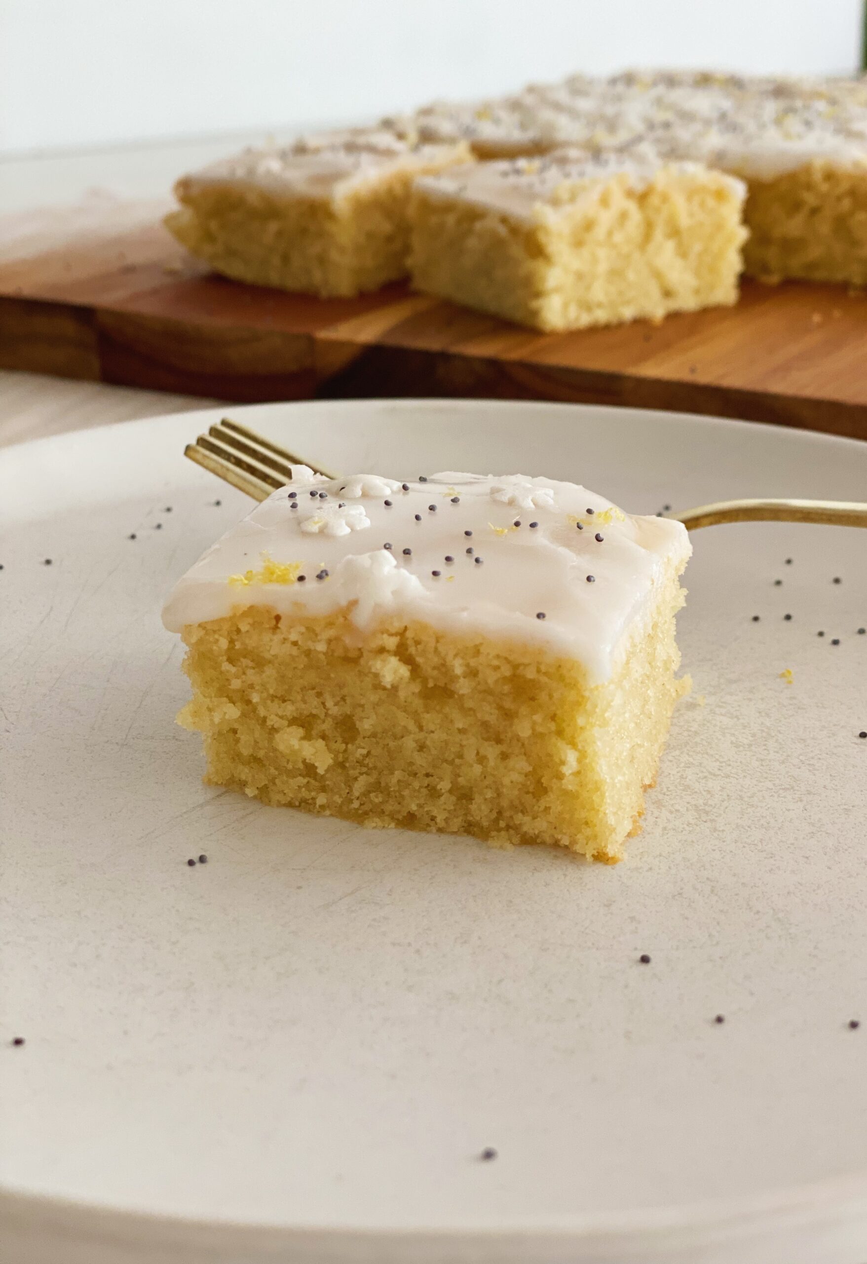 Et stykke citronkage på en hvid tallerken med en guld gaffel. I baggrunden er der et spækbræt med mere kage på.