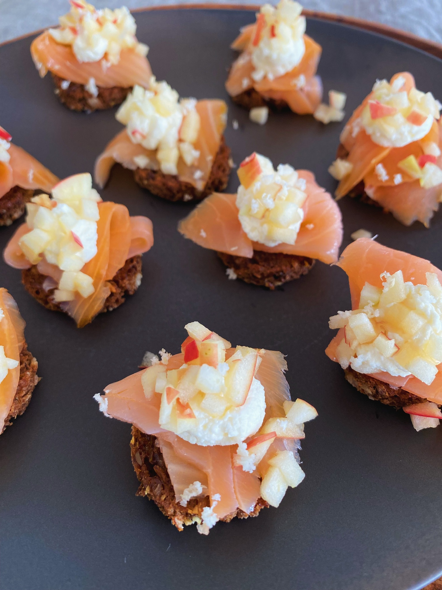 Rugbrødshapsere med røget laks og peberrodsfløde set skråt ind, hvor de bagerste er sløret. På en sort tallerken som står på et træfad. Det står på en hørdug.