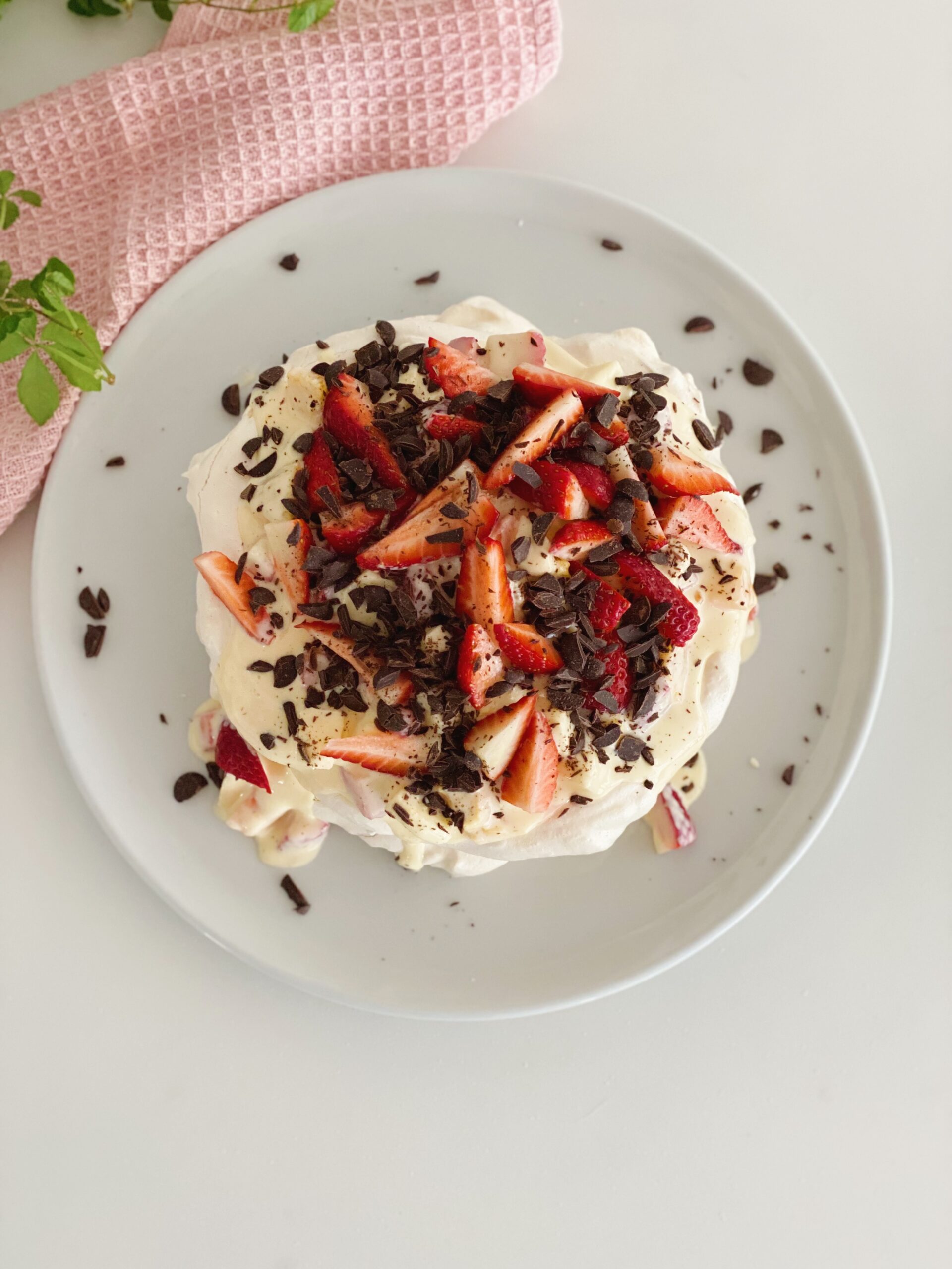 Marengslagkage med frugtsalat set tæt oppefra. Den står på et hvidt fad og der er en lyserød klud og nogle grønne blade i den ene side af billedet.