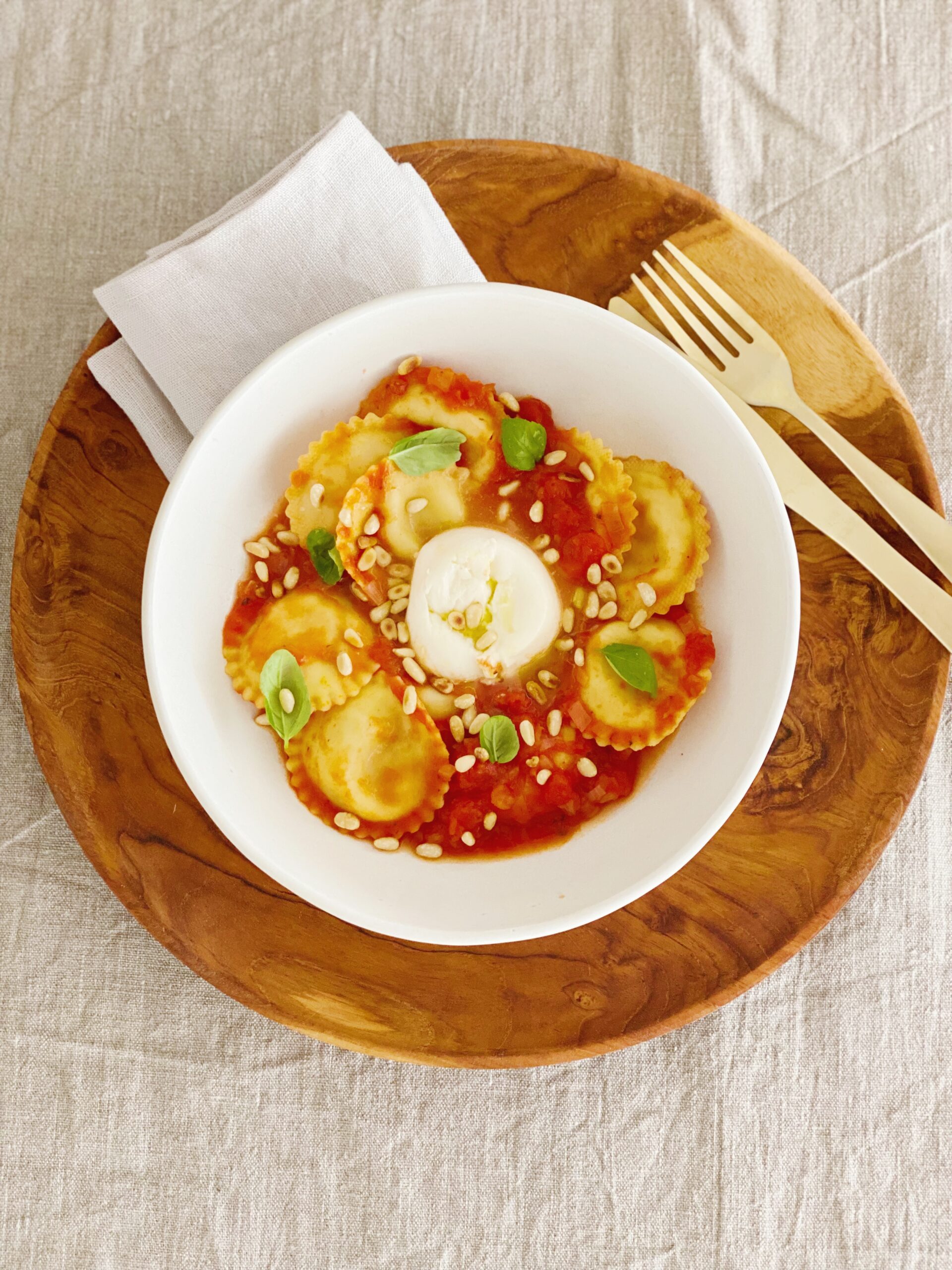 Ravioli med tomat og burrata set oppefra. Serveret i en hvid skål som står på et træfad. Ved siden af ligger der en kniv og gaffel