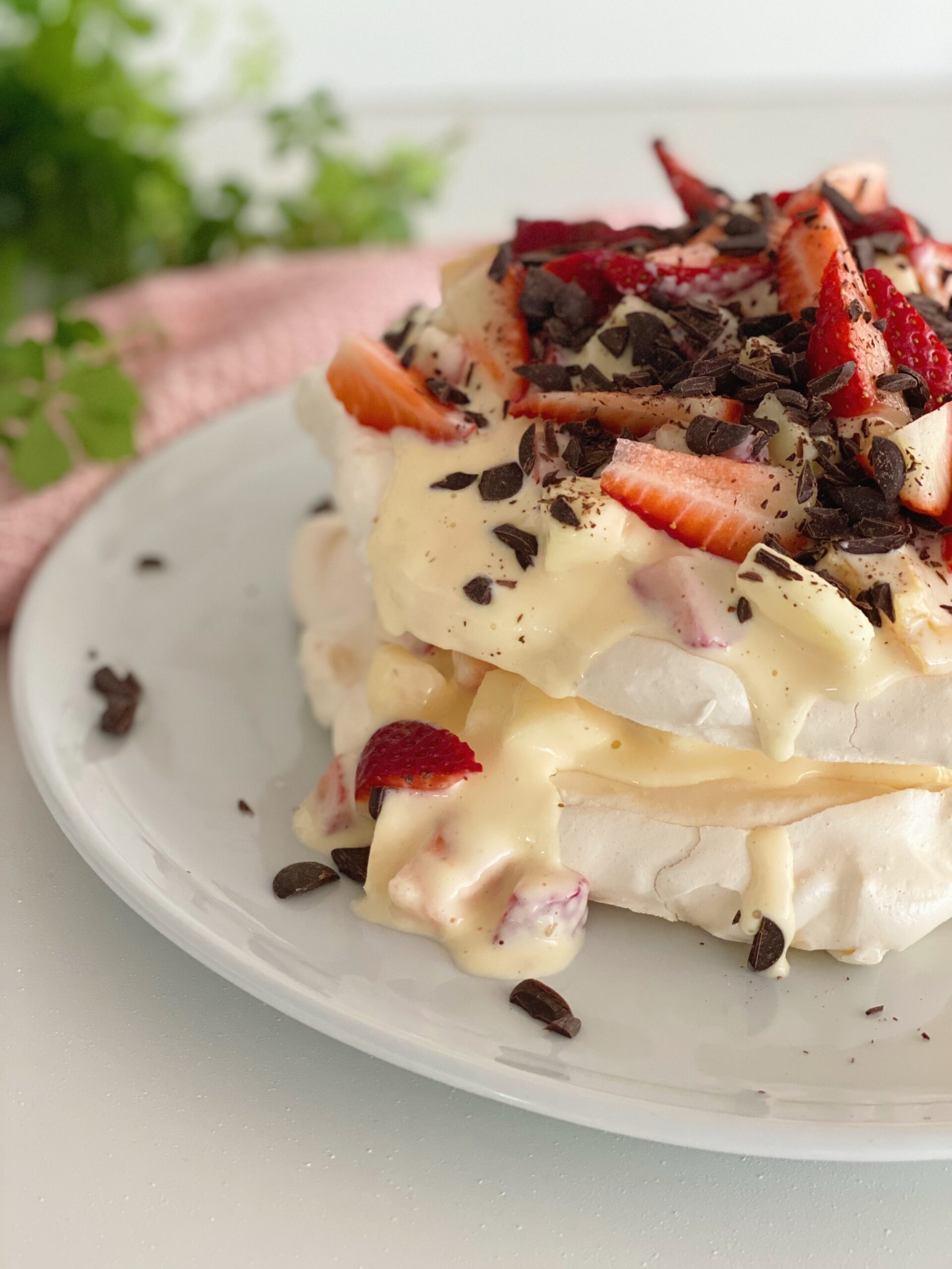 Marengslagkage med frugtsalat set tæt på, hvor du kan se halvdelen af lagkagen. Den står på et hvidt fad og der er en lyserød klud og nogle grønne blade i baggrunden
