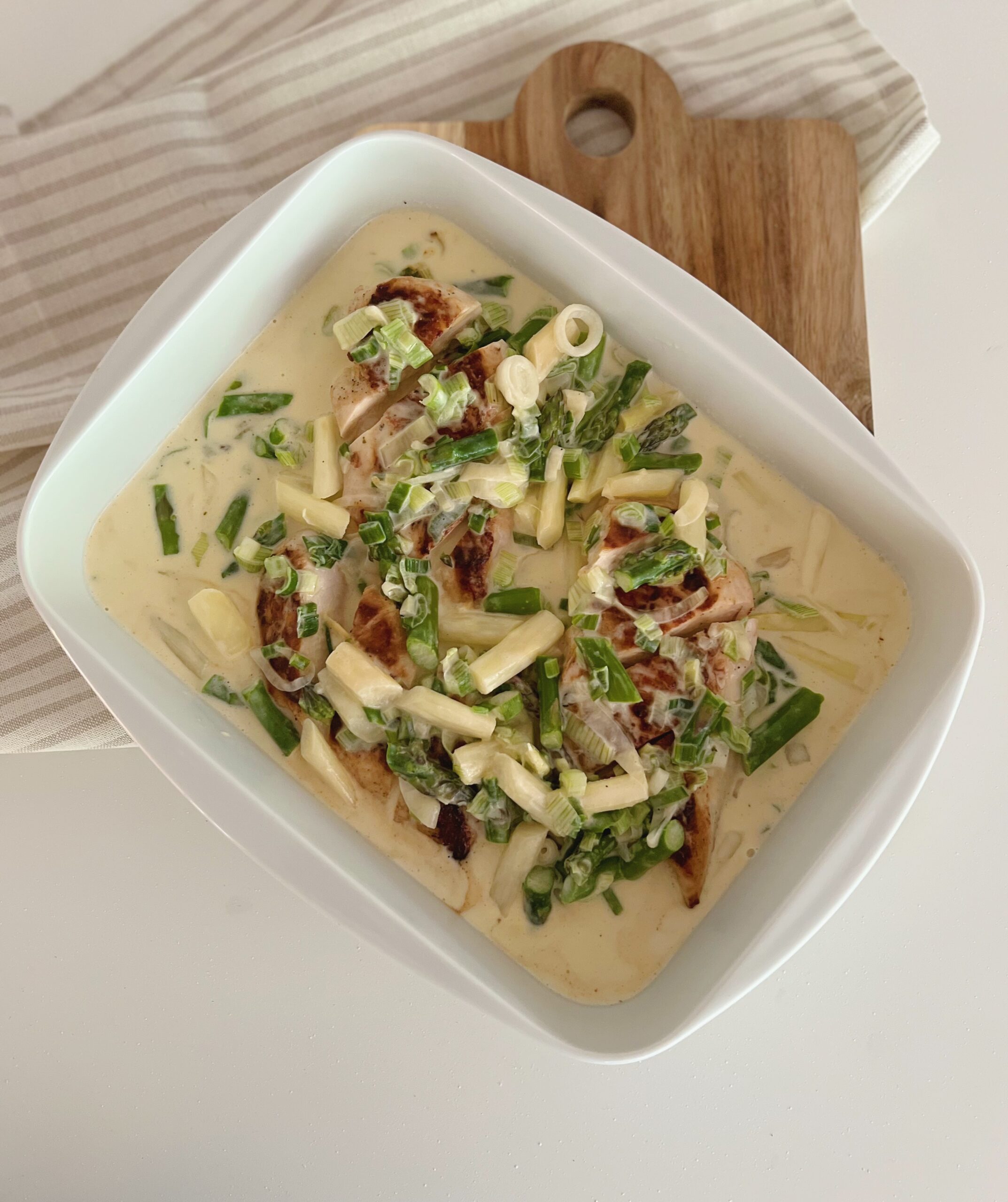 Kylling i cremet sauce med asparges i et firkantet fad, som står oven på et træspækbræt med en stribet stof serviet under. Fadet står skråt på spækbrættet.