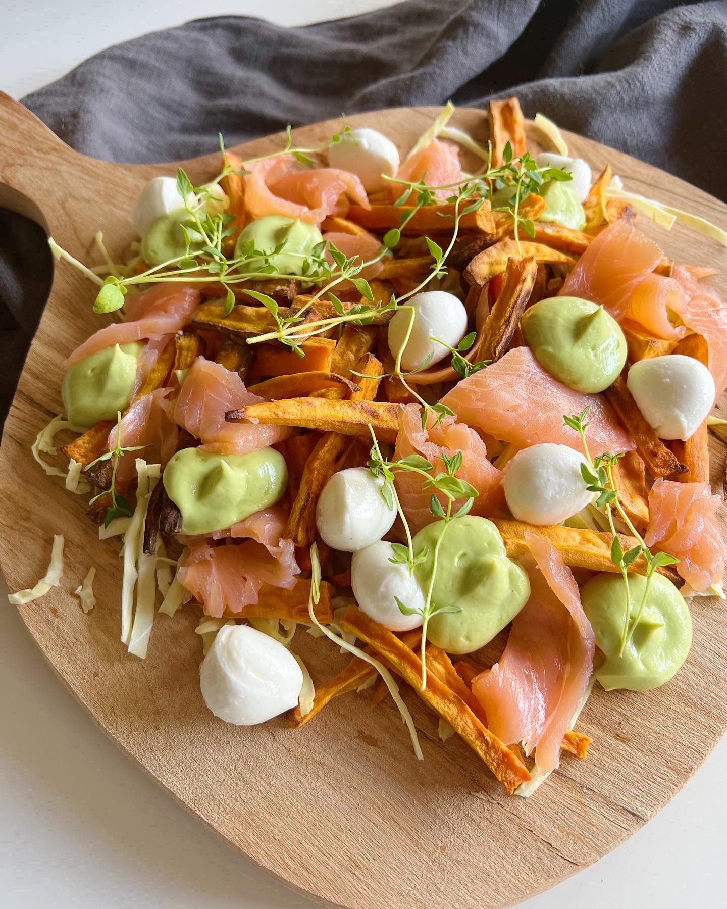 Sprød laksesalat med søde kartofler og avocadocreme serveret på en planke/spækbræt. Set tæt på, på et hvidt bord med en mørke grå klud bagved