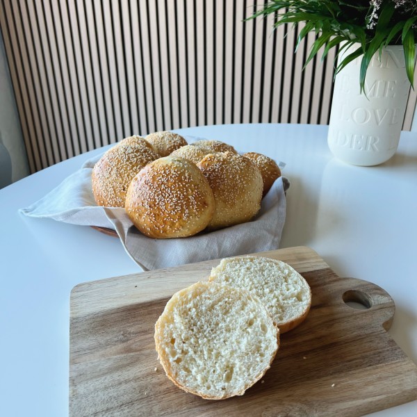 Burgerboller med danskvand på et fad med en grå stofserviet i bunden. Foran er der en af bollerne som er skåret over så man kan se den indeni. Det hele står på et hvidt bord med en vase ved siden af hvor du kan se bladende fra en buket og en akustik væg i baggrunden.