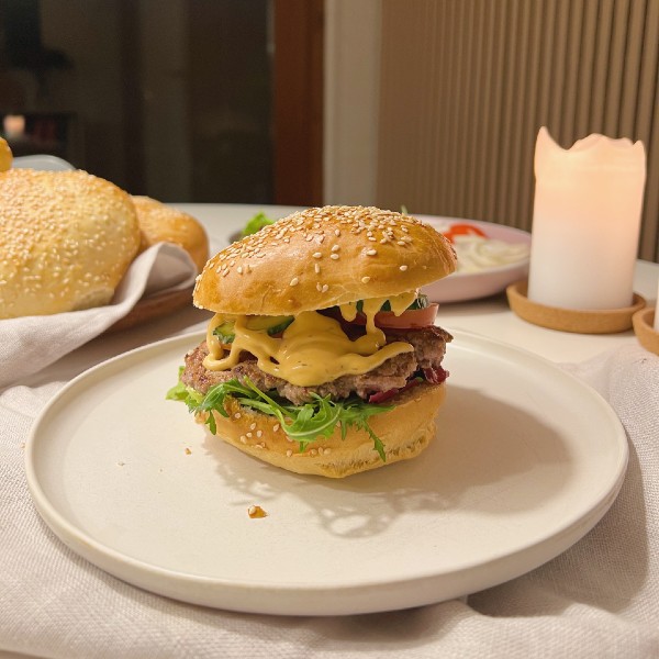 Her ser du en hjemmelavet burger med salat i bunden, en bøf, agurk, tomat og dressing. I baggrunden kan man se flere burgerboller på et fad og en tallerken med tilbehør på og et starinlys der er tændt. Baggrunden er en akustik væg