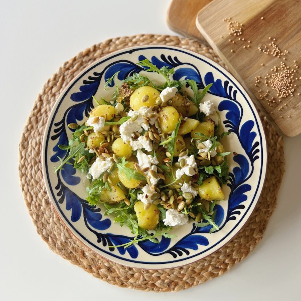 Brasede kartofler med rucola og feta set oppefra på et græsk inspireret fad i blå og hvide farver. Ved siden af fadet er der et spækbræt i træ hvor der ligger sennepsfrø