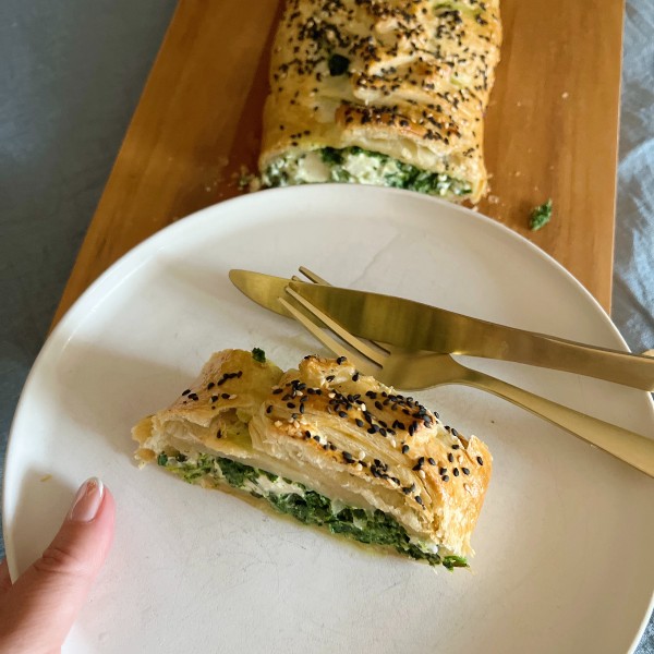 En spanakopita stang ses i baggrunden og i forgrunden ser du en hvid tallerken med et stykke på. Der ligger en guld gaffel og kniv på tallerkenen