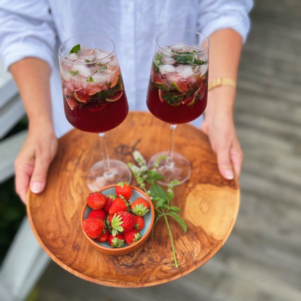 To jordbær mojitos på et rundt træfad. Der er også en skål med jordbær, og en stilk med mynte på bakken. Bakken bliver holdt af et menneske med hvid skjorte. Man kan kun se armene og lidt af overkroppen