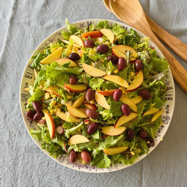 sommersalat med nektariner og grillede majs set oppefra. salaten er på et rundt fad og der ligger et salatsæt i træ ved siden af